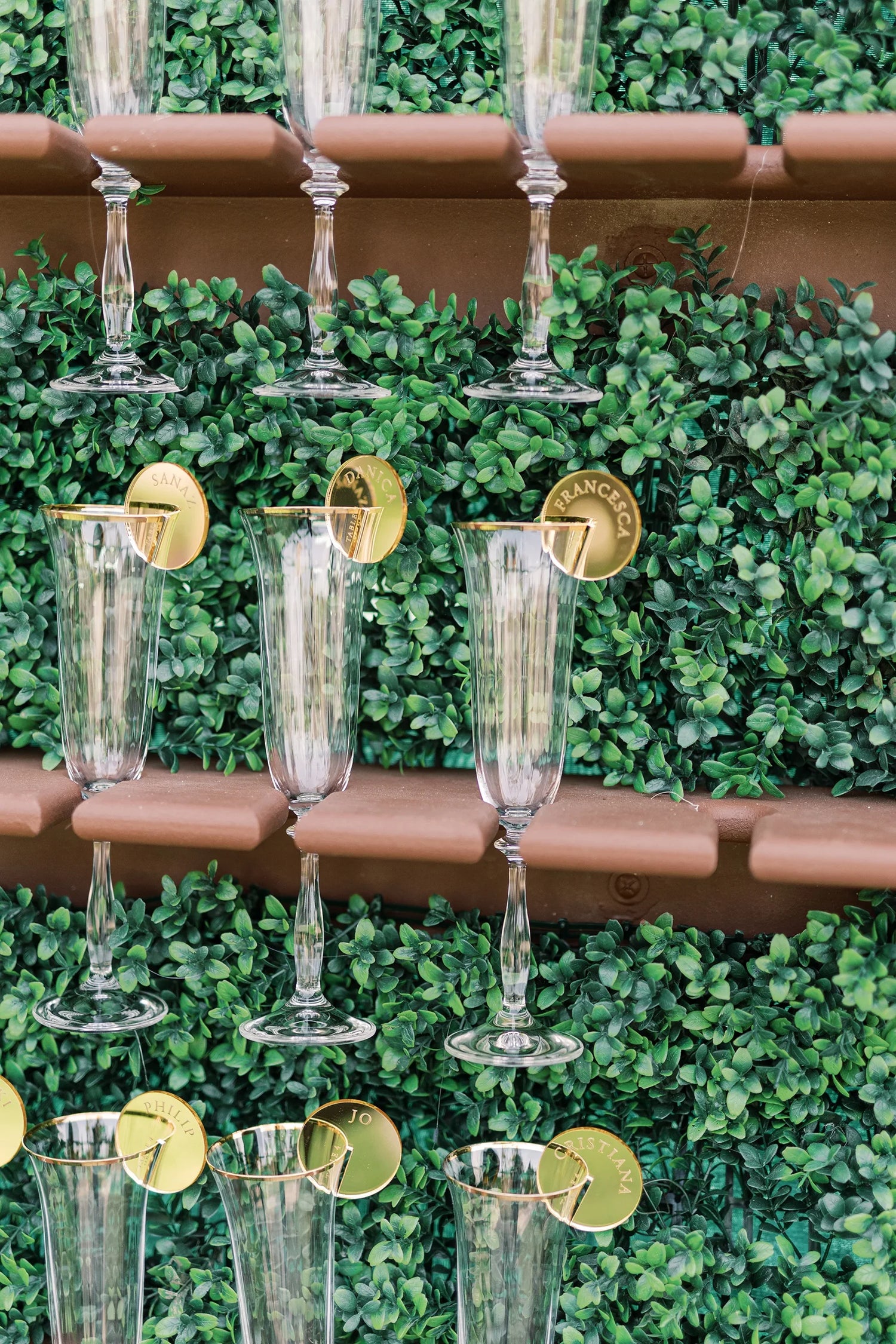 Stylish champagne display with clear glasses neatly arranged on a lush green foliage wall, each adorned with personalised gold acrylic name tags.