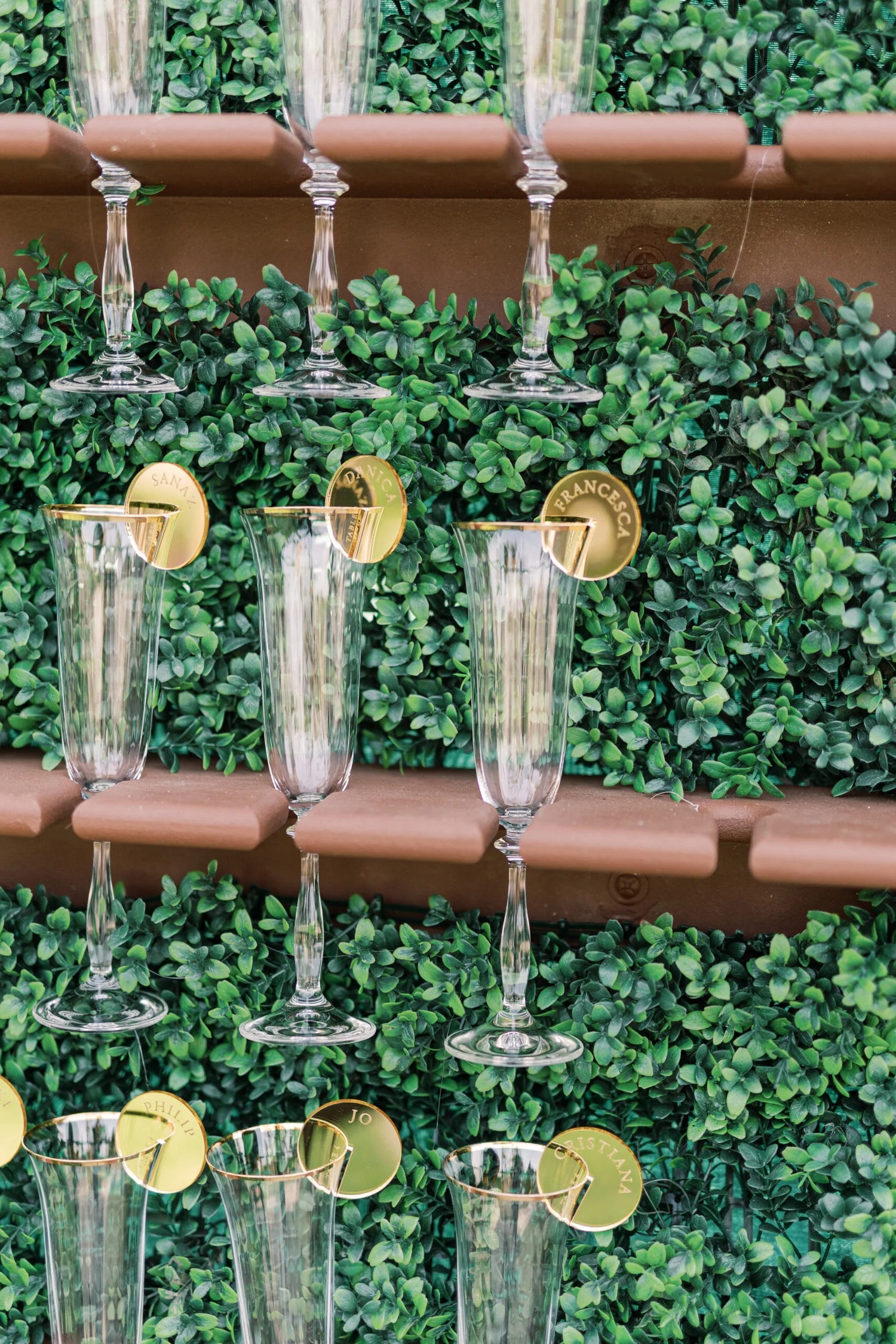 Stylish champagne display with clear glasses arranged on a lush green wall, each adorned with personalized gold acrylic name tags.