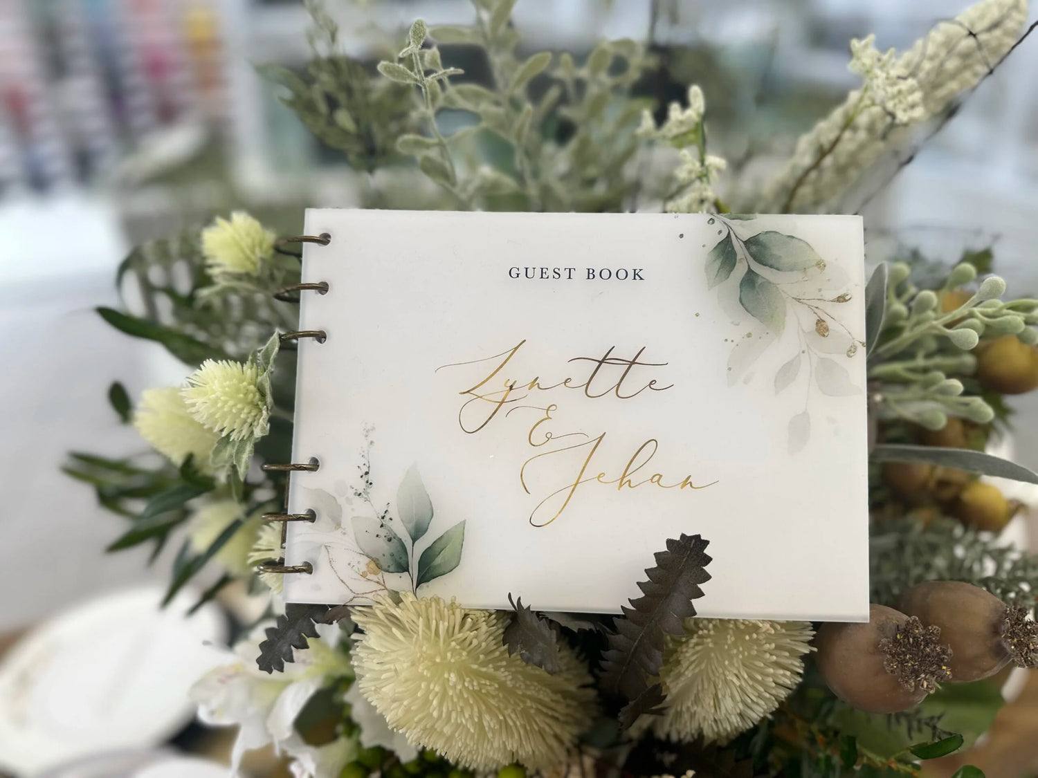 Botanical-themed wedding guest book for Lynette and Jehan, featuring green eucalyptus illustrations, gold foil lettering, and a floral arrangement backdrop.