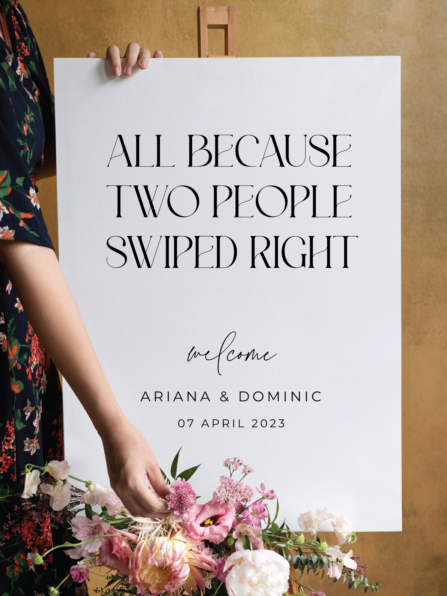 A playful white wedding welcome sign that reads “All Because Two People Swiped Right” in bold black serif font. The couple's names, Ariana and Dominic, along with the wedding date, are displayed below. A floral arrangement with pink, white, and green blooms decorates the easel.
