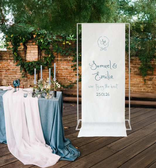Elegant fabric wedding welcome sign with Samuel & Emilia's names, set beside a romantic blue and white table arrangement outdoors.
