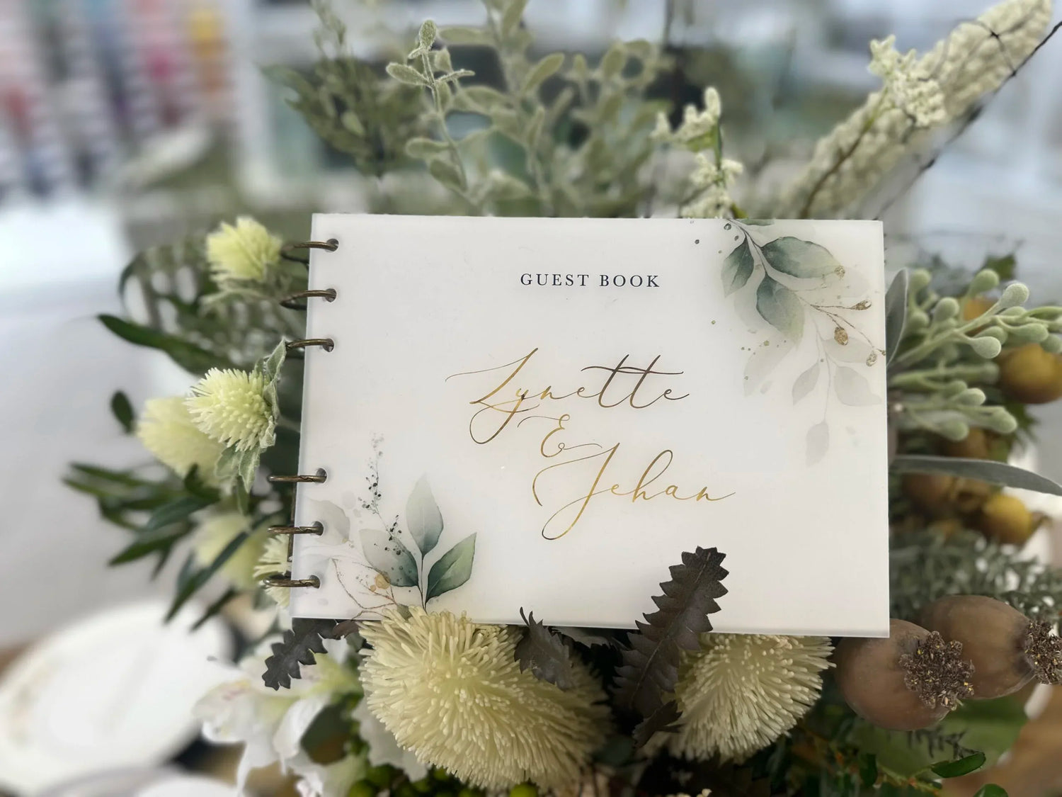 Elegant acrylic guest book featuring Lynette & Jehan’s names in gold calligraphy, surrounded by soft white and green floral arrangements.