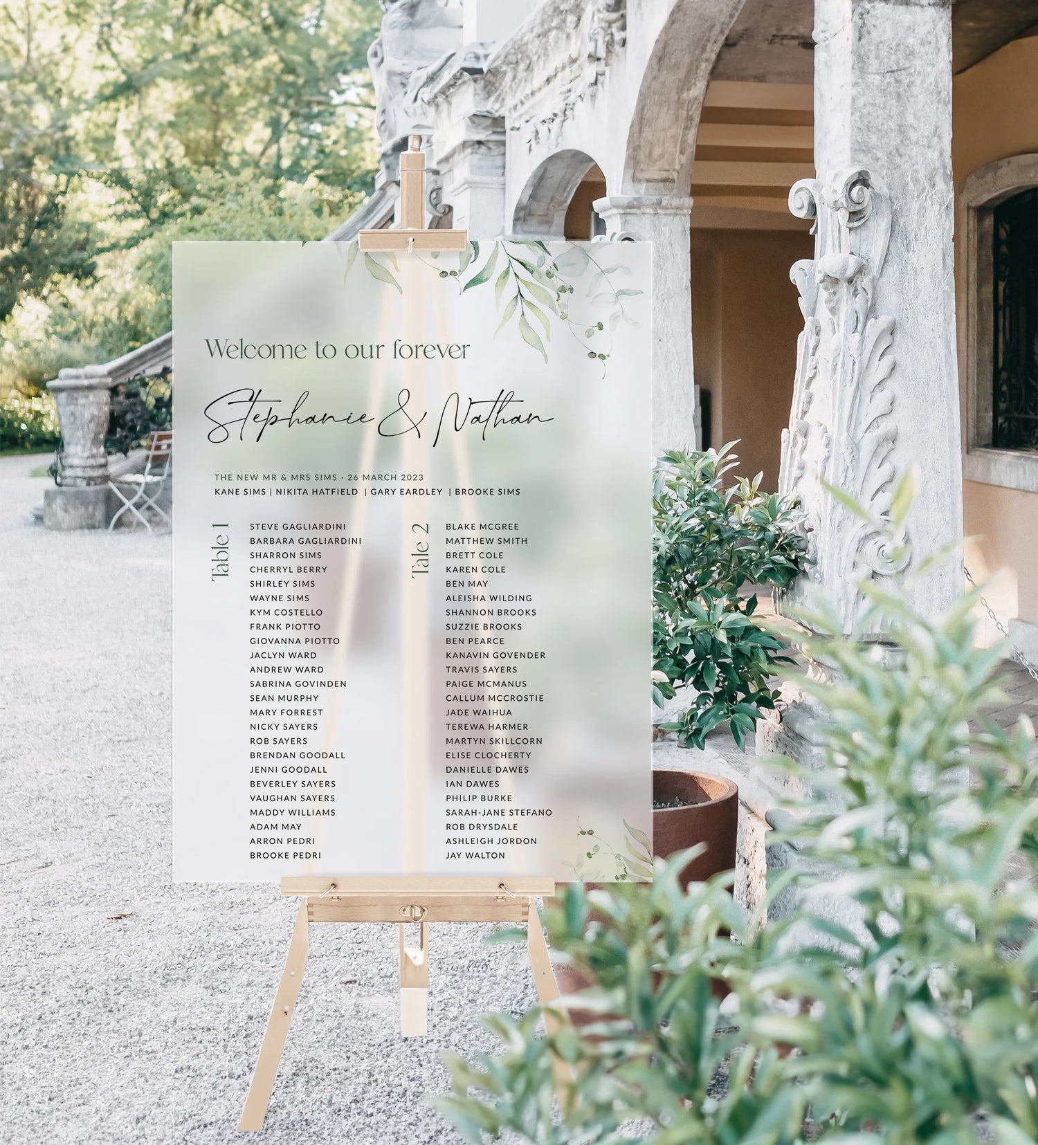 An elegant acrylic wedding seating chart for Stephanie and Nathan, displayed on a wooden easel outdoors. The sign features a soft green botanical design with table assignments listed in two columns. A classical stone building and lush greenery provide a scenic backdrop.