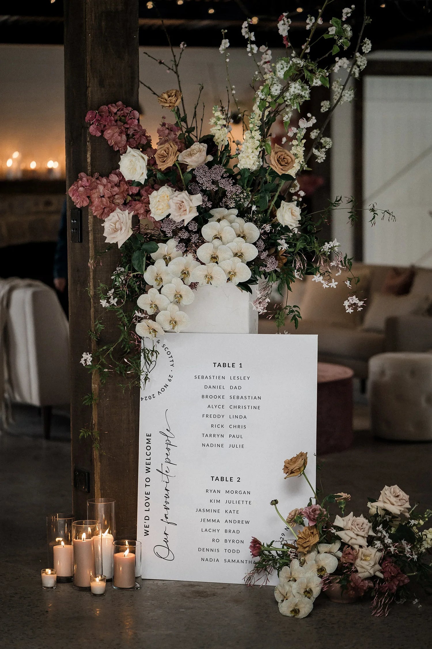 A romantic wedding seating chart displayed on a white board, adorned with lush floral arrangements of roses and wildflowers. The chart is placed against a rustic wooden beam, surrounded by glowing candles, creating an intimate and elegant ambiance. A cosy reception area with soft lighting and stylish furniture is visible in the background.