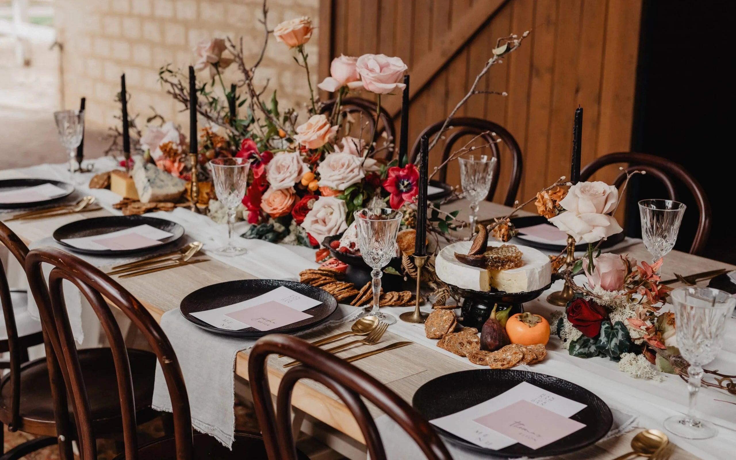 Rustic wedding table setting with dark plates, menus and place cards, gold cutlery, crystal glassware, and a centrepiece featuring vibrant roses, candles, and a cheese and fruit display