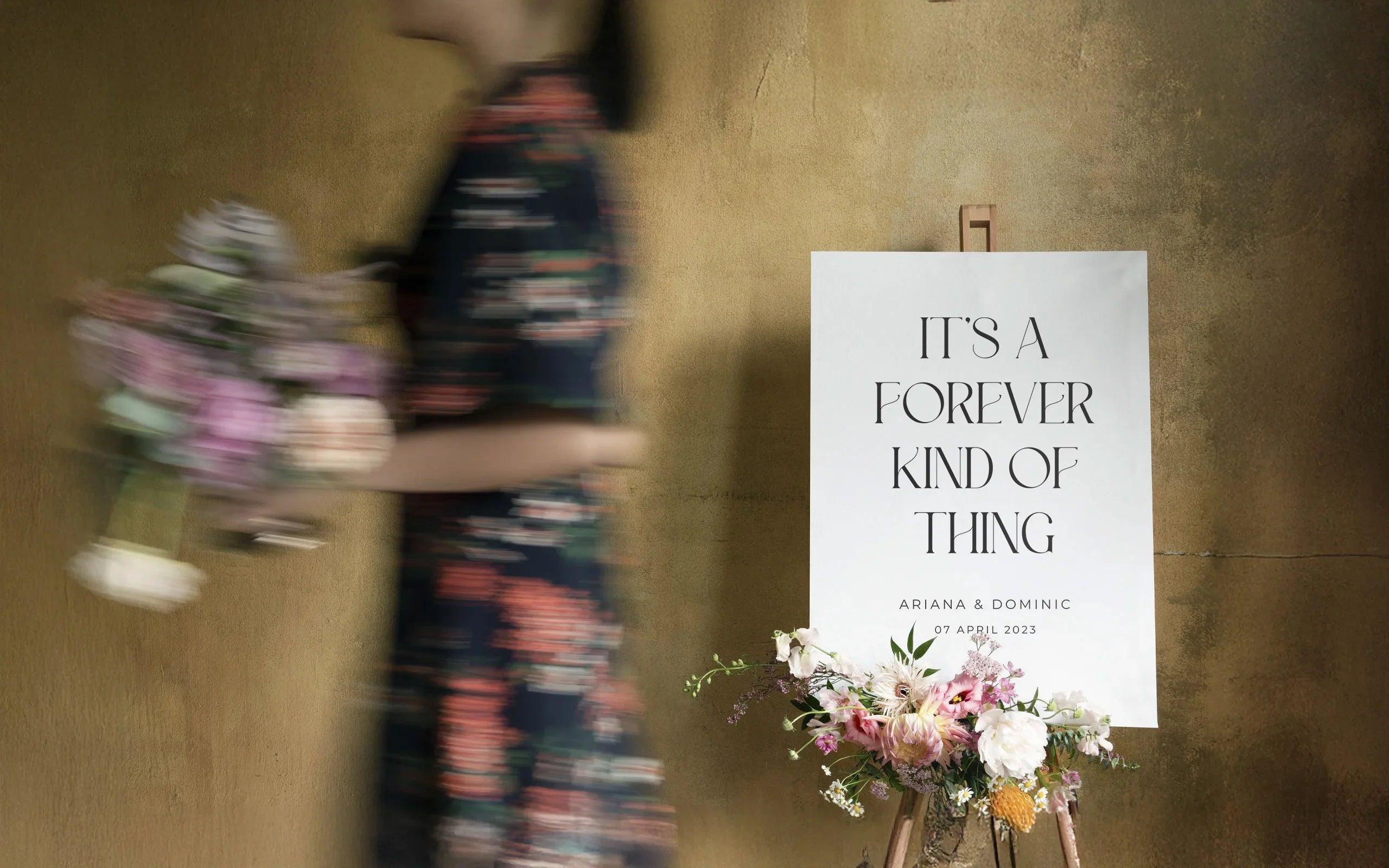 A wedding sign on an easel with 'It's a Forever Kind of Thing' text, surrounded by floral arrangements, and a blurred figure holding a bouquet passing by in the background.