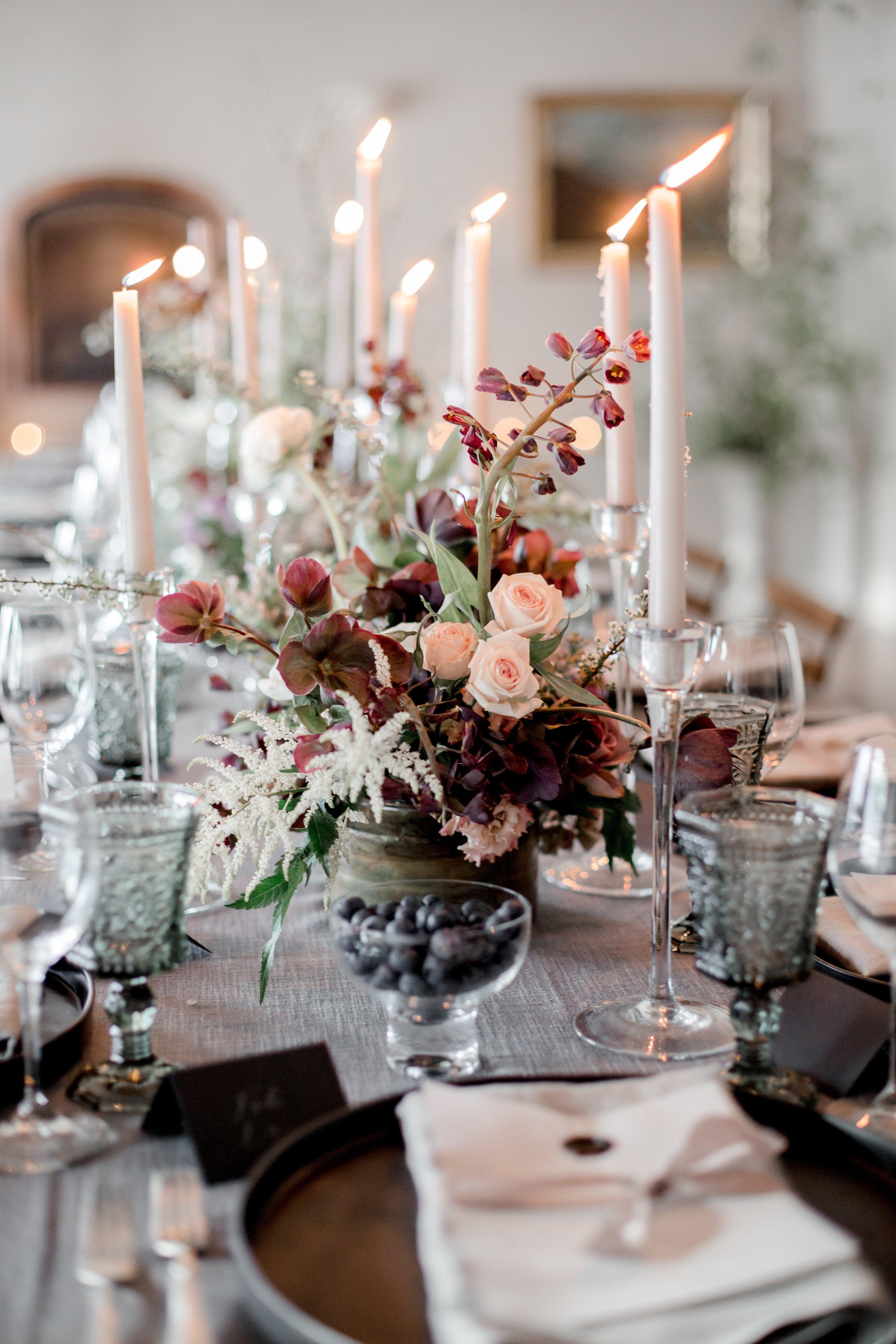 Elegant wedding table decor featuring a lush floral centrepiece in deep hues, surrounded by tall white taper candles and vintage glassware
