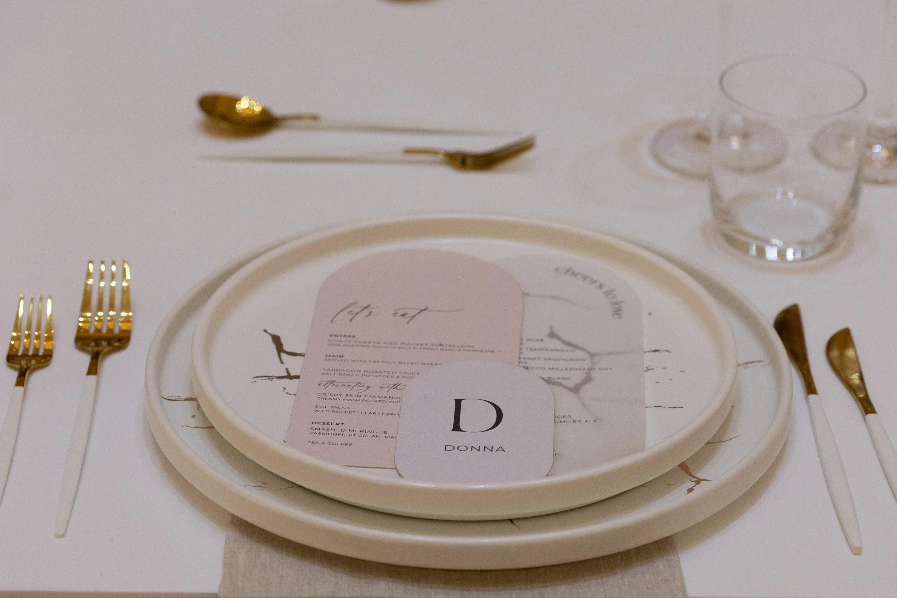 Minimalist wedding table setting with a double arch place card featuring the guest's name and an elegant blush arch-shaped menu on a marble dinner plate