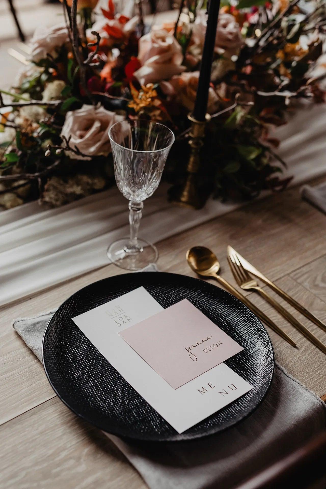 Black textured dinner plate featuring a modern minimalist wedding menu with a personalized square guest name card for Joanne Elton, styled with gold cutlery and a floral centerpiece.