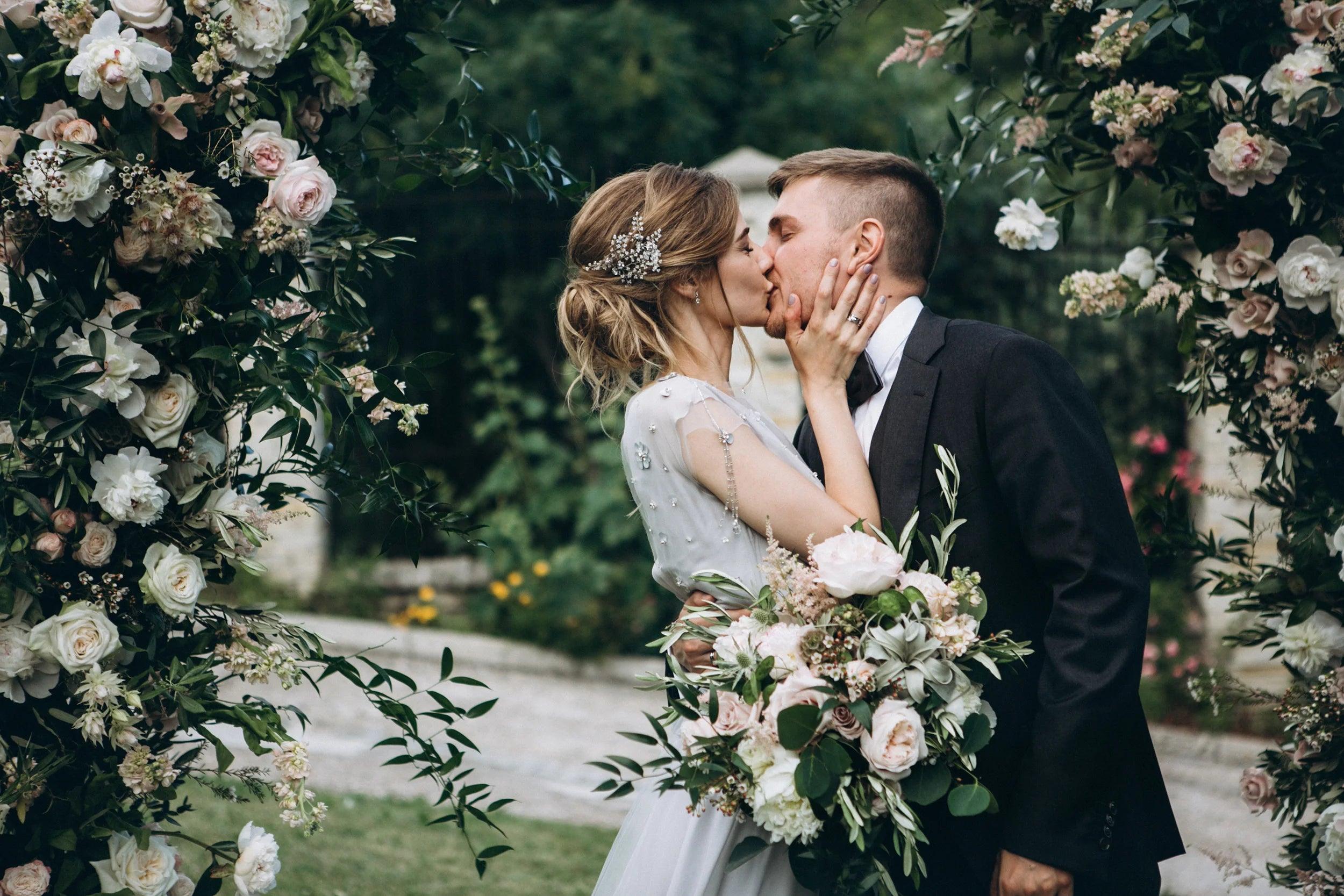 Bride and groom kissing under a floral wedding arch, surrounded by lush greenery and romantic blooms