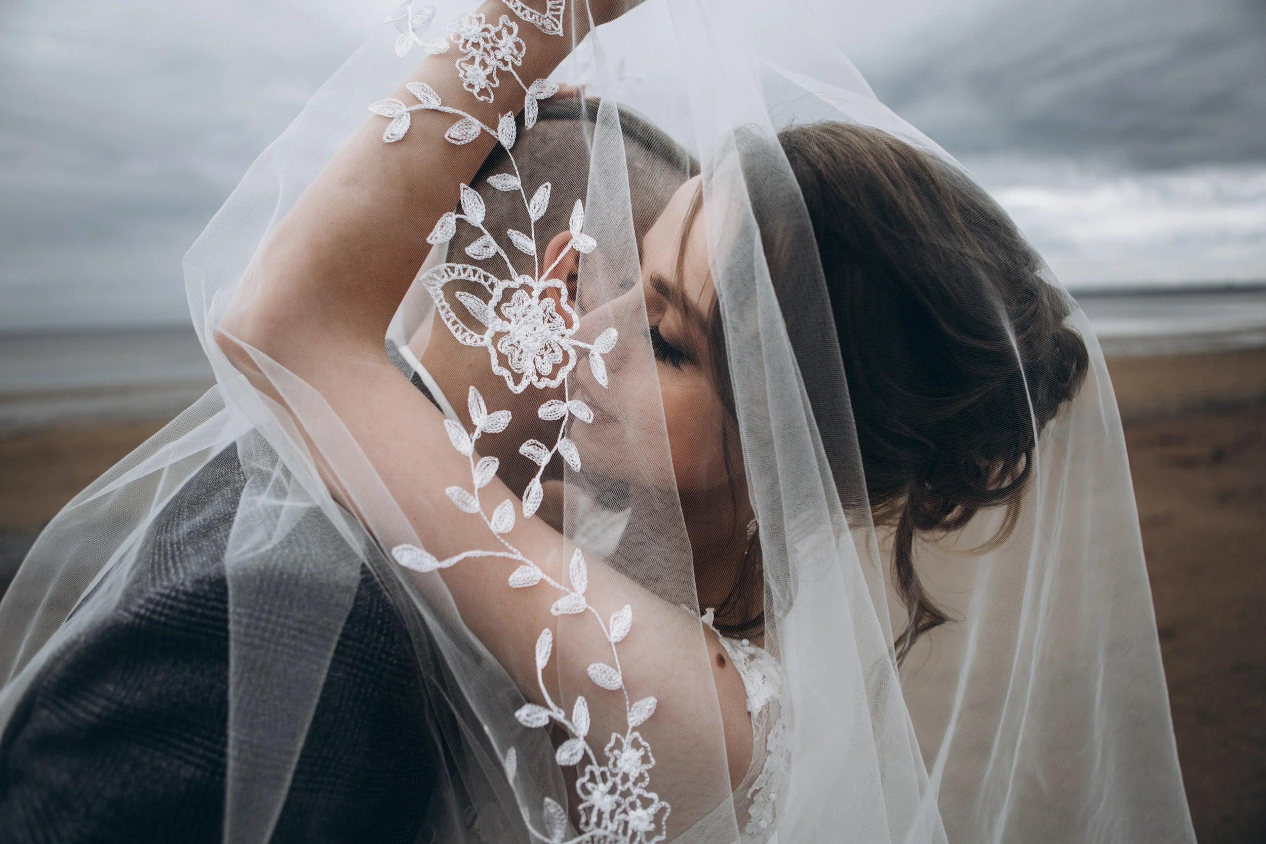 Bride and groom share a romantic kiss under a delicate lace wedding veil on a windy beach.