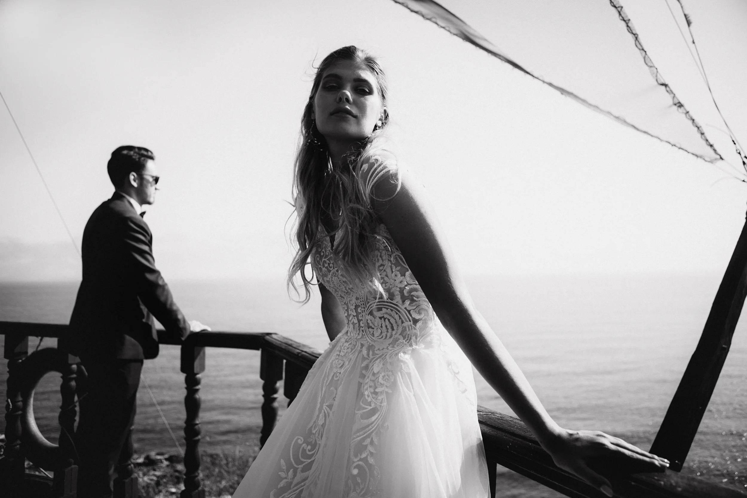 Black-and-white wedding photo of a bride in a lace gown leaning on a railing, with the groom in the background overlooking the ocean.
