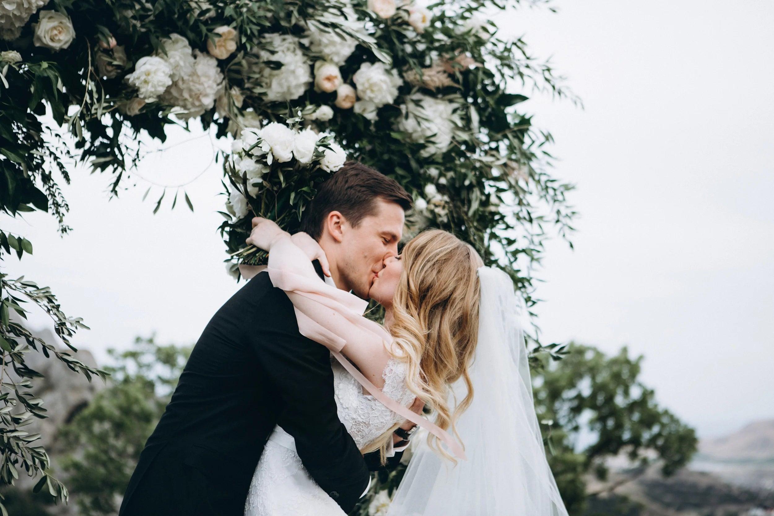 Bride and groom sharing a romantic kiss under a floral wedding arch, celebrating their special day outdoors