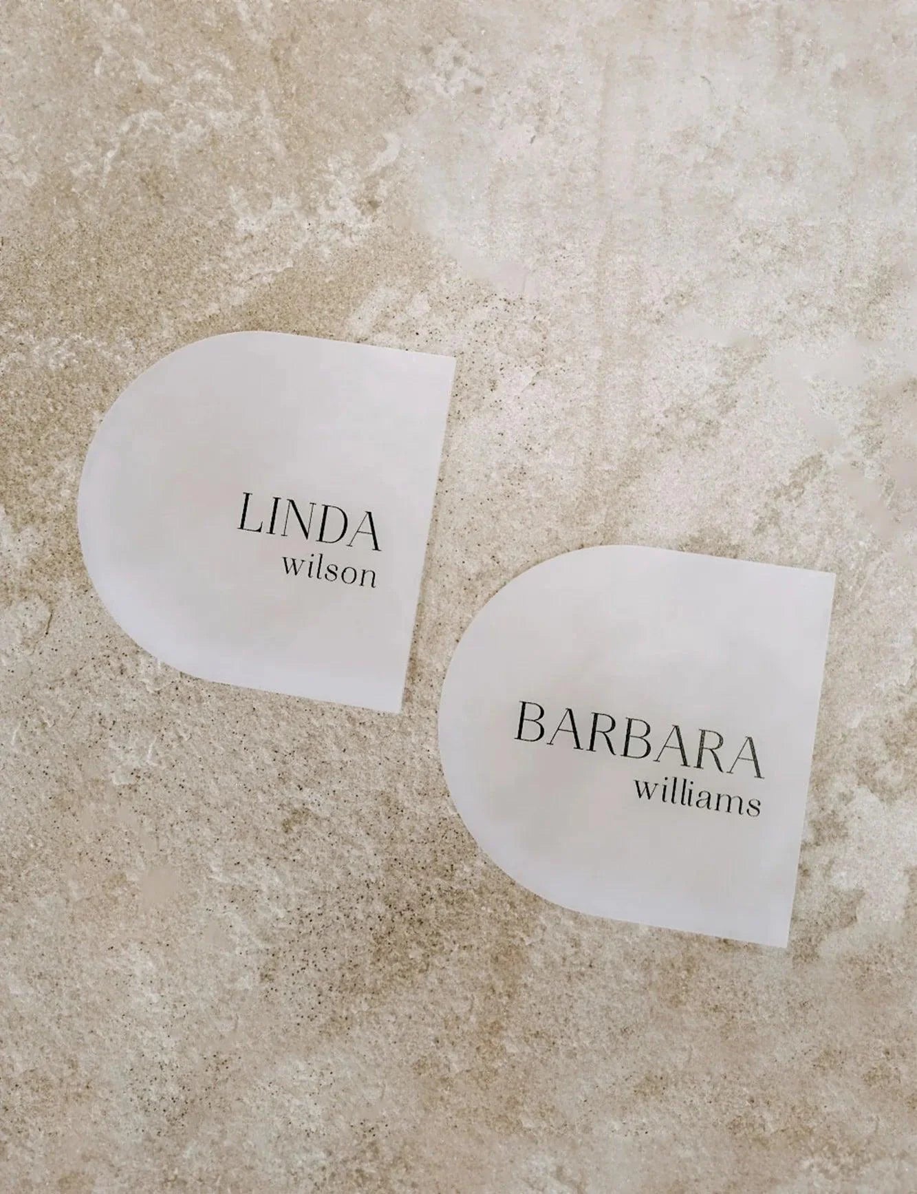 Sophisticated arched acrylic coasters, place cards displaying names Linda Wilson and Barbara Williams, photographed on a textured stone surface.