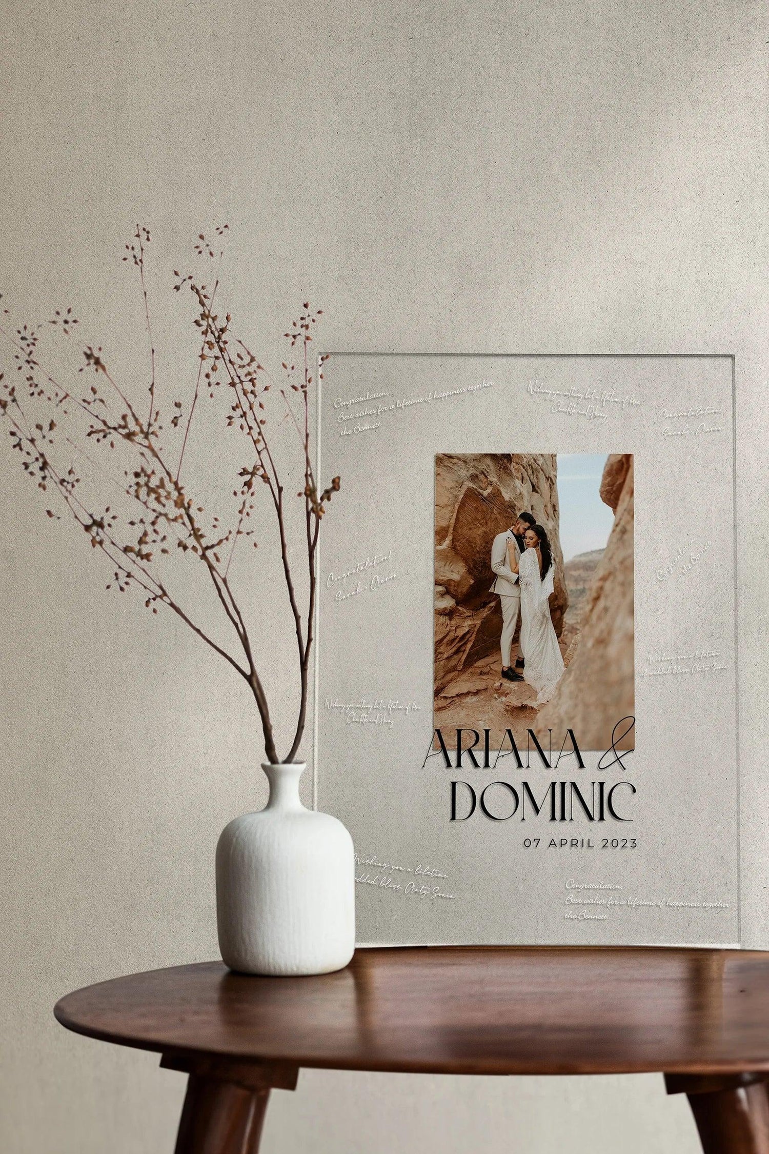 A modern acrylic photo sign featuring Ariana & Dominic, dated 07 April 2023, with handwritten guest messages etched around the edges, propped against a neutral wall.