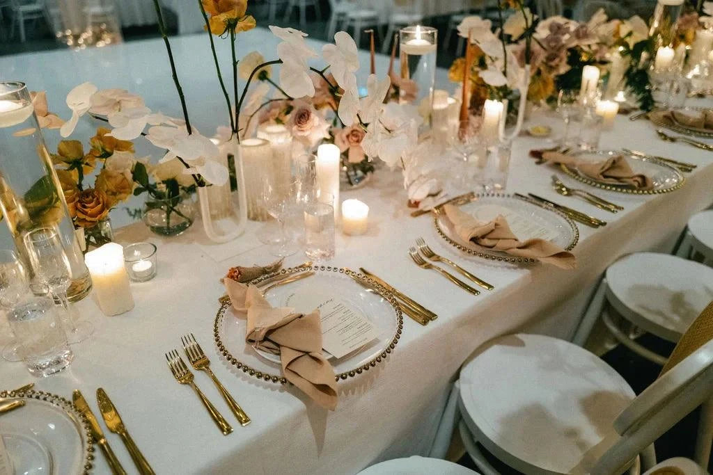 A luxurious wedding reception table styled with white tablecloths, gold cutlery, clear beaded charger plates, and soft candlelight, surrounded by floral arrangements of orchids and roses.