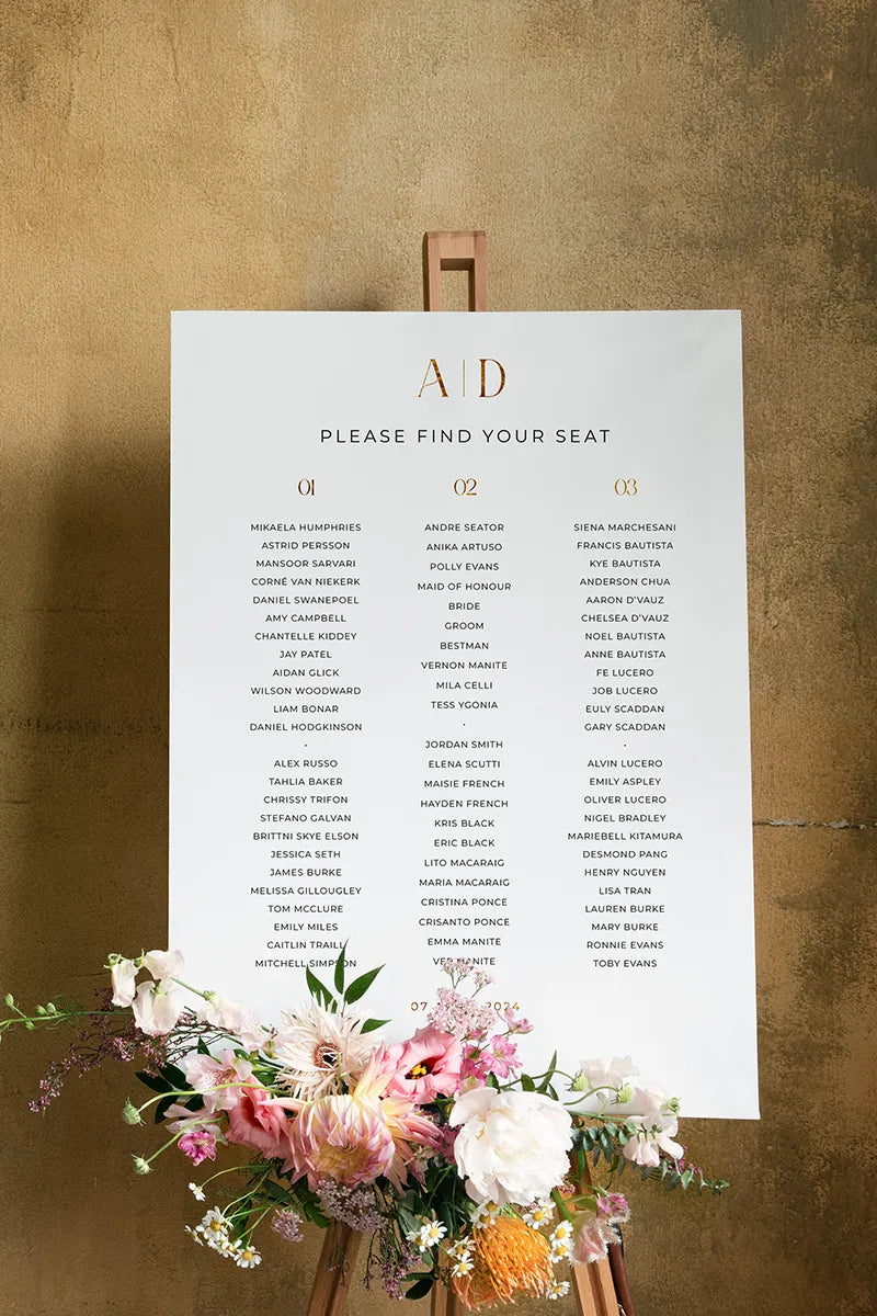 Elegant seating plan displayed on a gold easel with floral arrangements at the base, featuring guest names organized into table numbers.