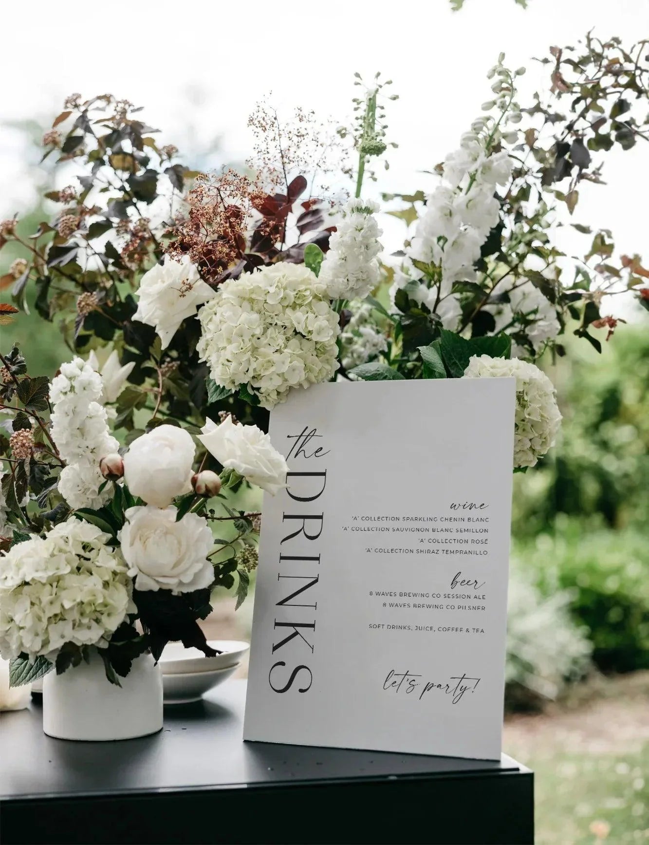 Modern acrylic bar sign featuring a stylish drinks menu with clean typography, surrounded by lush white floral arrangements on a black bar surface at an outdoor venue.