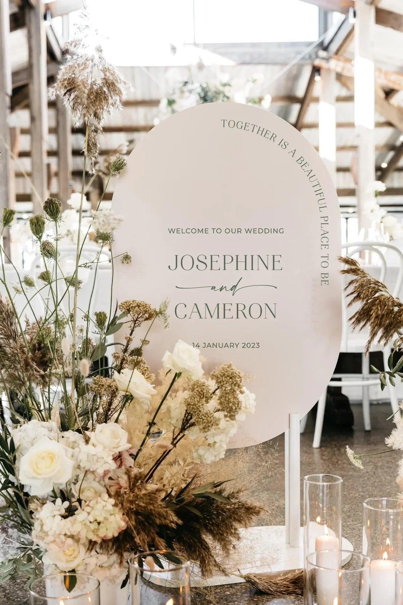 Double-arched wedding welcome sign for Josephine and Cameron with elegant green and black typography, surrounded by neutral florals in a rustic indoor venue setting.