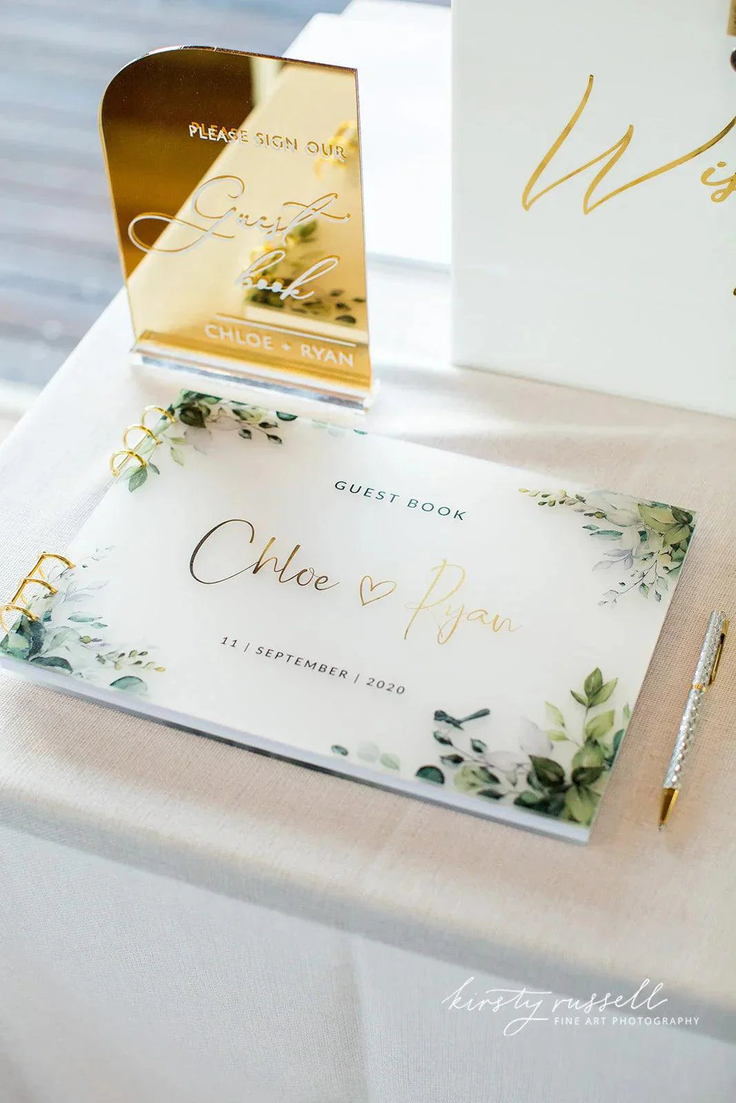 An acrylic guest book with Chloe & Ryan in gold script, surrounded by greenery illustrations, placed on a wedding table with a gold mirrored Please Sign Our Guest Book sign.