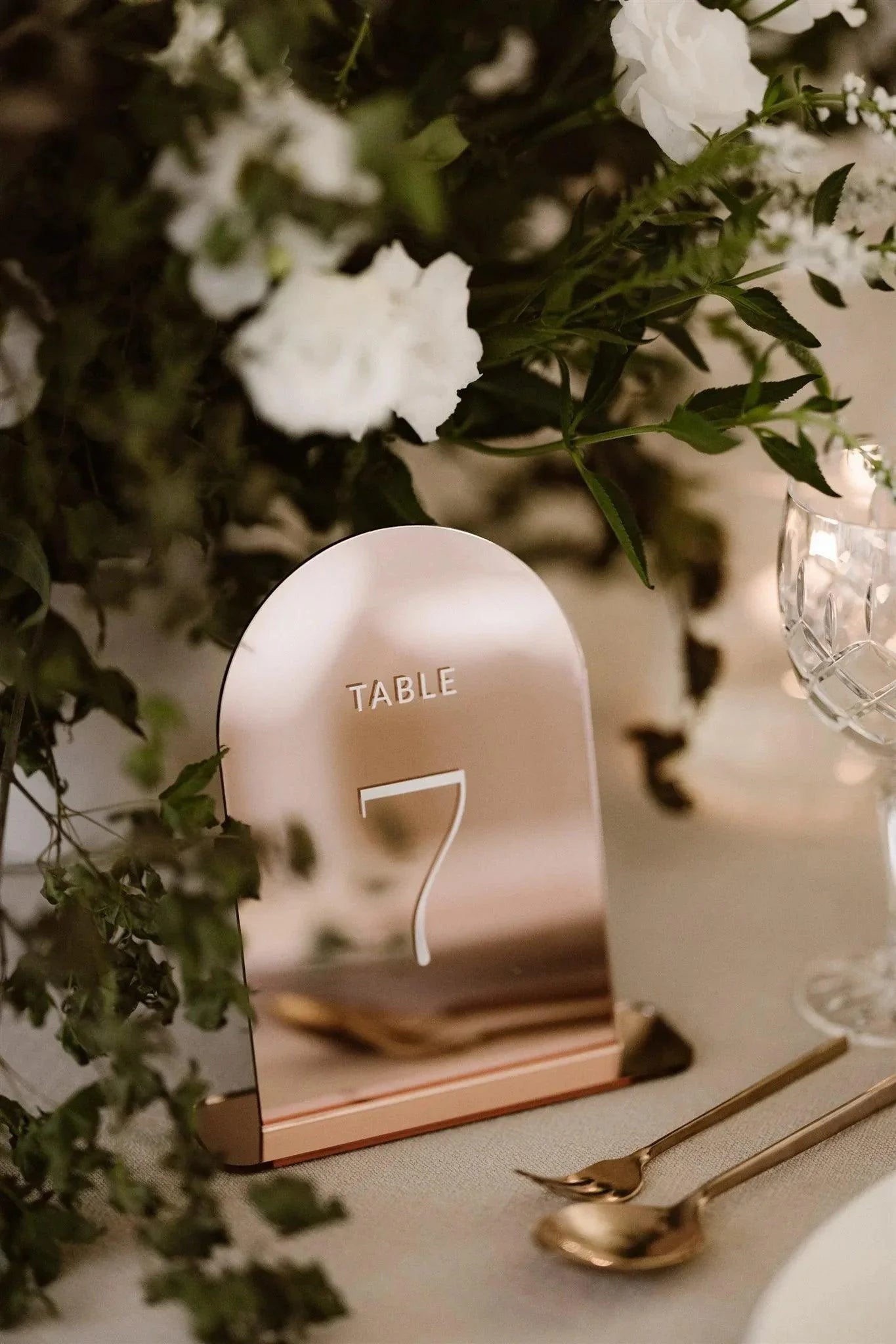 Modern rose gold acrylic table number with Table 7 displayed on a wedding table surrounded by lush greenery and white flowers.