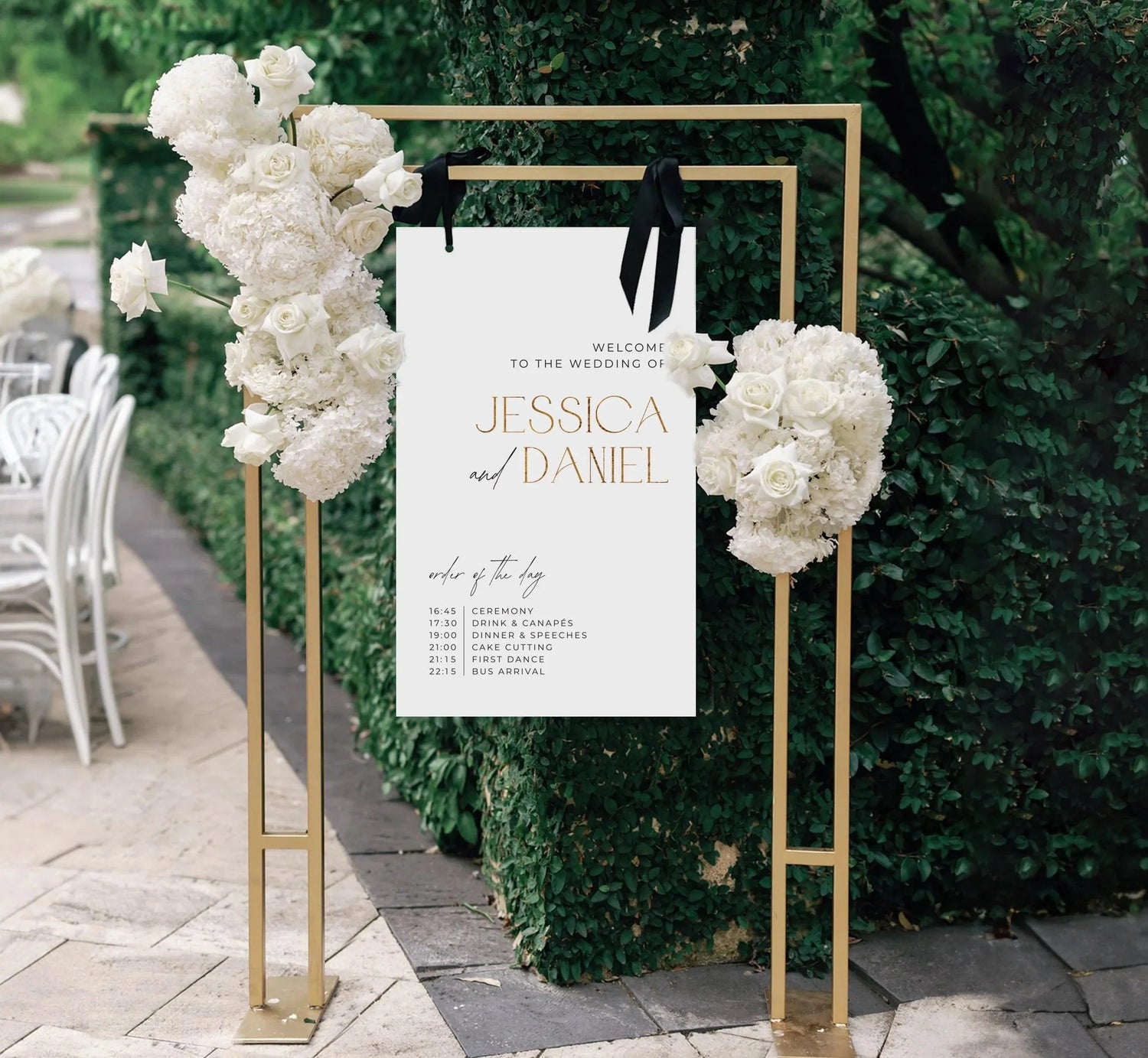 Modern white wedding welcome sign for Jessica and Daniel, decorated with lush white flowers on a gold stand, placed outdoors.