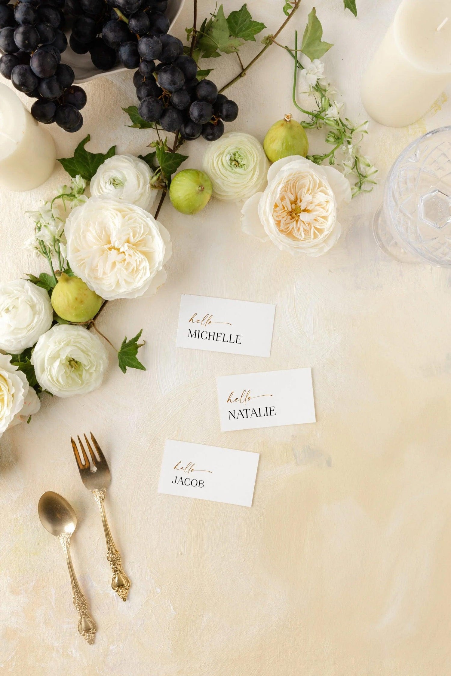 Elegant name place cards reading Michelle, Natalie, and Jacob styled on a cream-colored table with fresh figs, grapes, white roses, and vintage gold flatware.