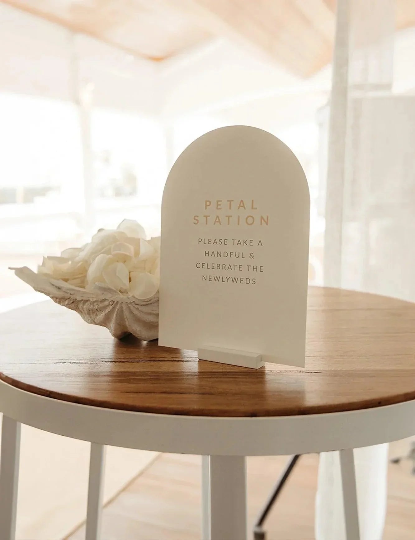 Minimalist, arch-shaped acrylic sign reading “Petal Station,” encouraging guests to celebrate the newlyweds, placed on a wooden table alongside a shell filled with white petals.