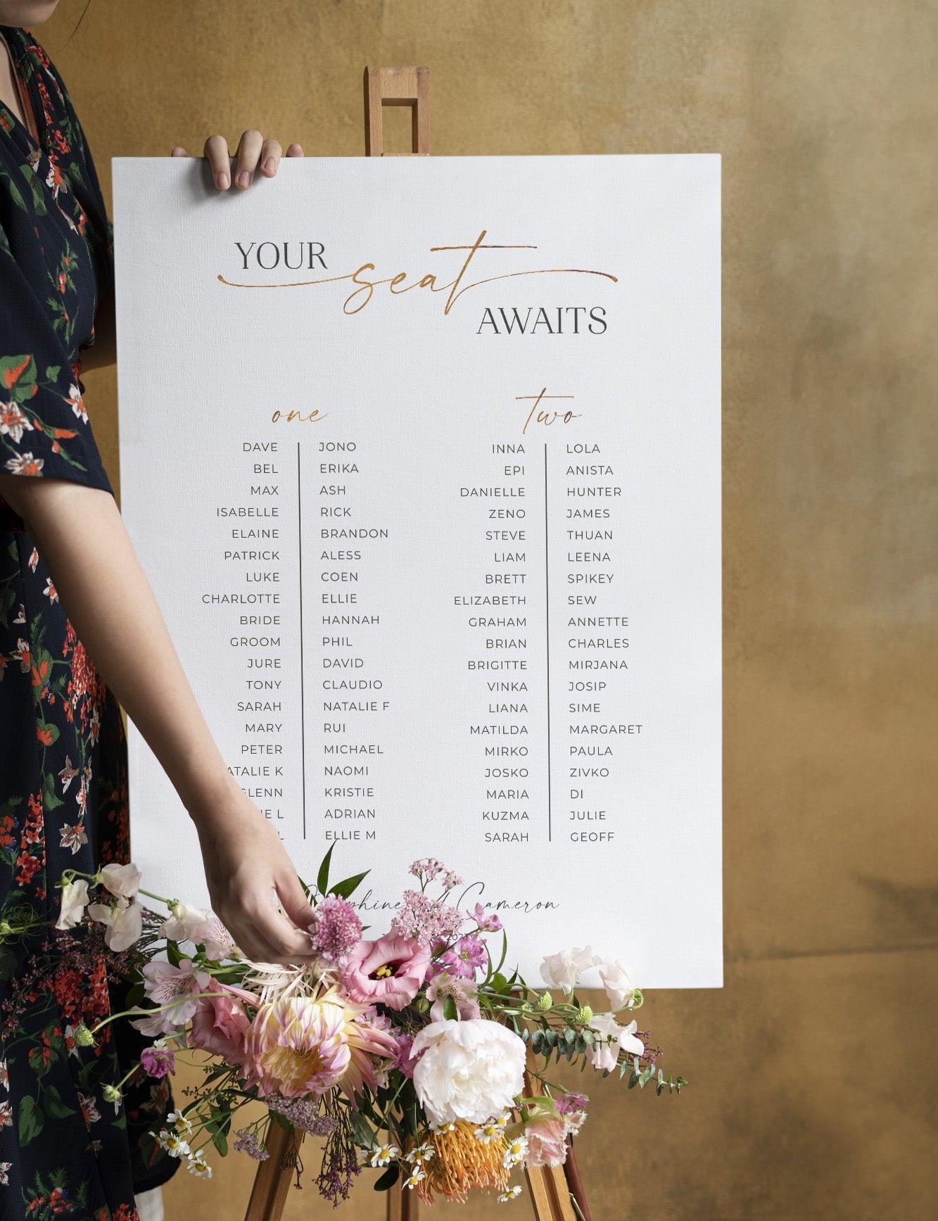 Stylish white seating chart with gold calligraphy, titled “Your Seat Awaits,” displayed on a wooden easel adorned with fresh florals, against a warm gold backdrop.