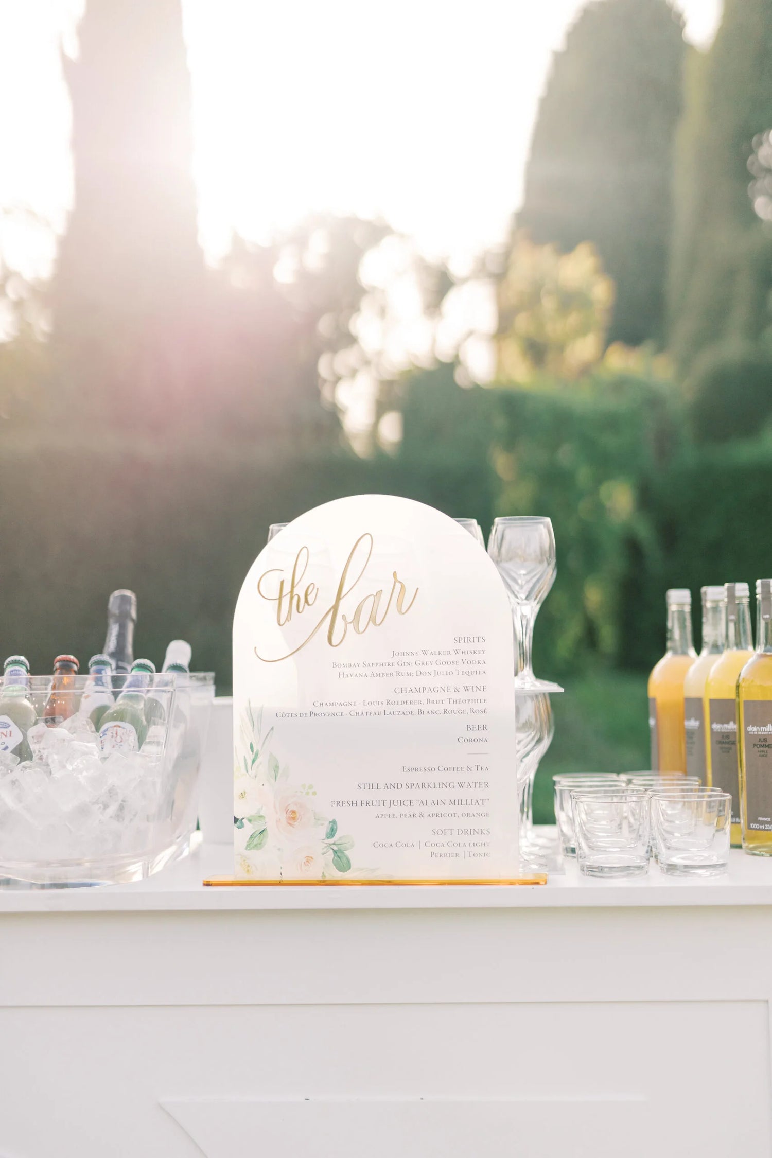 Elegant acrylic bar sign with gold calligraphy detailing the drinks menu, surrounded by glassware and bottled beverages on an outdoor bar with soft natural lighting.