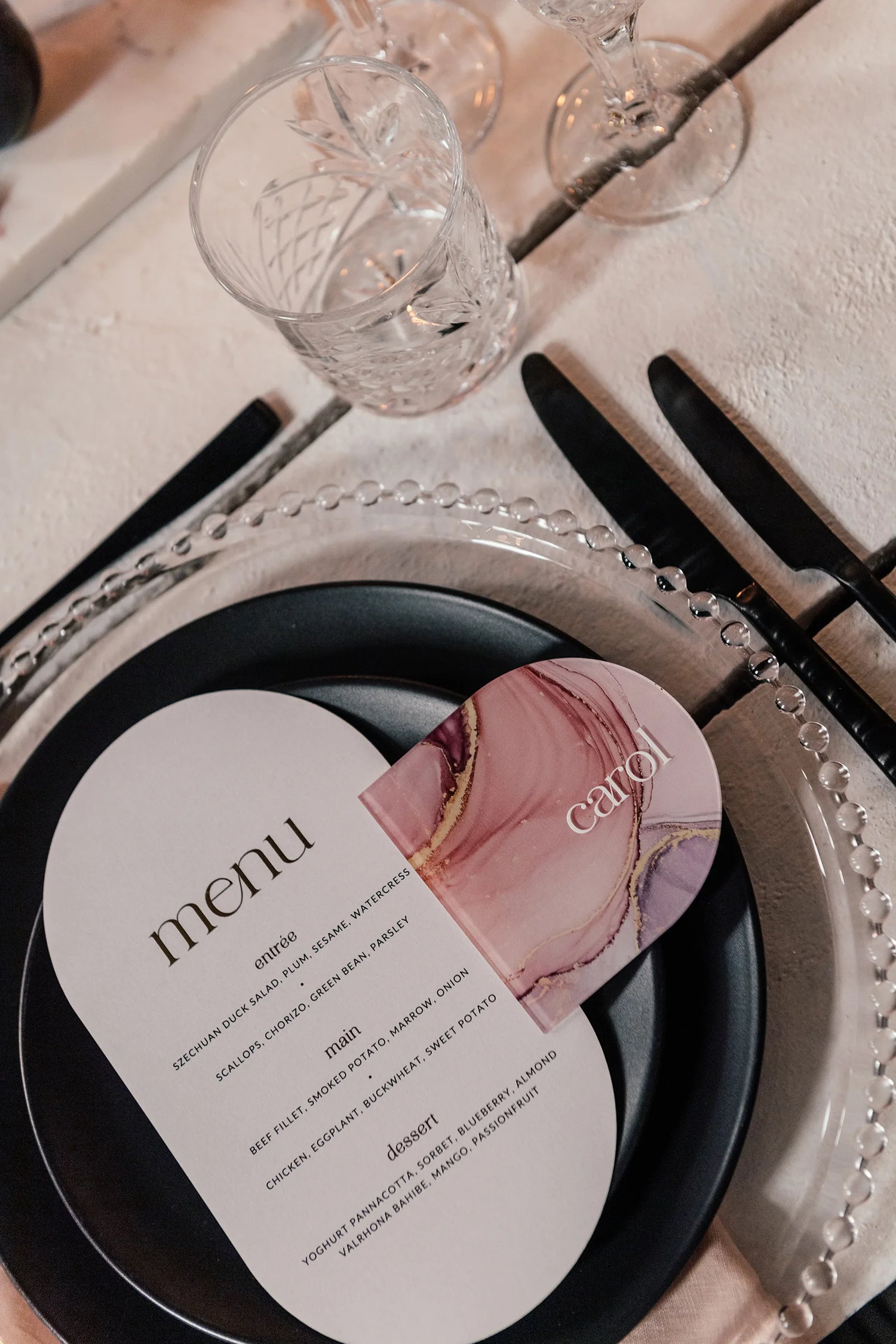 Modern wedding menu paired with a personalised acrylic coaster name card in pink marble design, elegantly displayed on a black plate with crystal glassware.