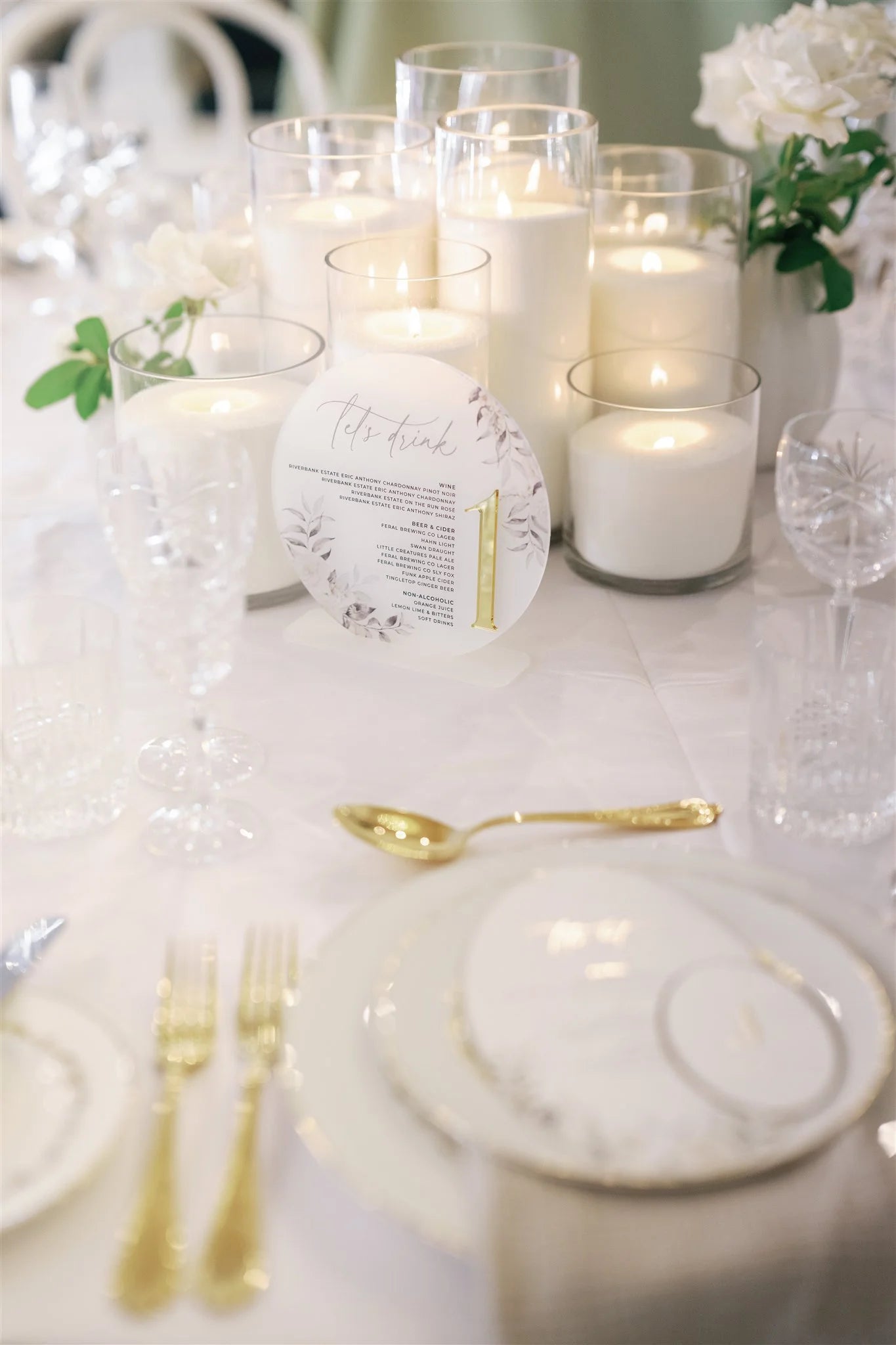 Elegant wedding table setting featuring an acrylic table number with gold accents, surrounded by tall white candles in glass holders, and a delicate floral arrangement in soft tones