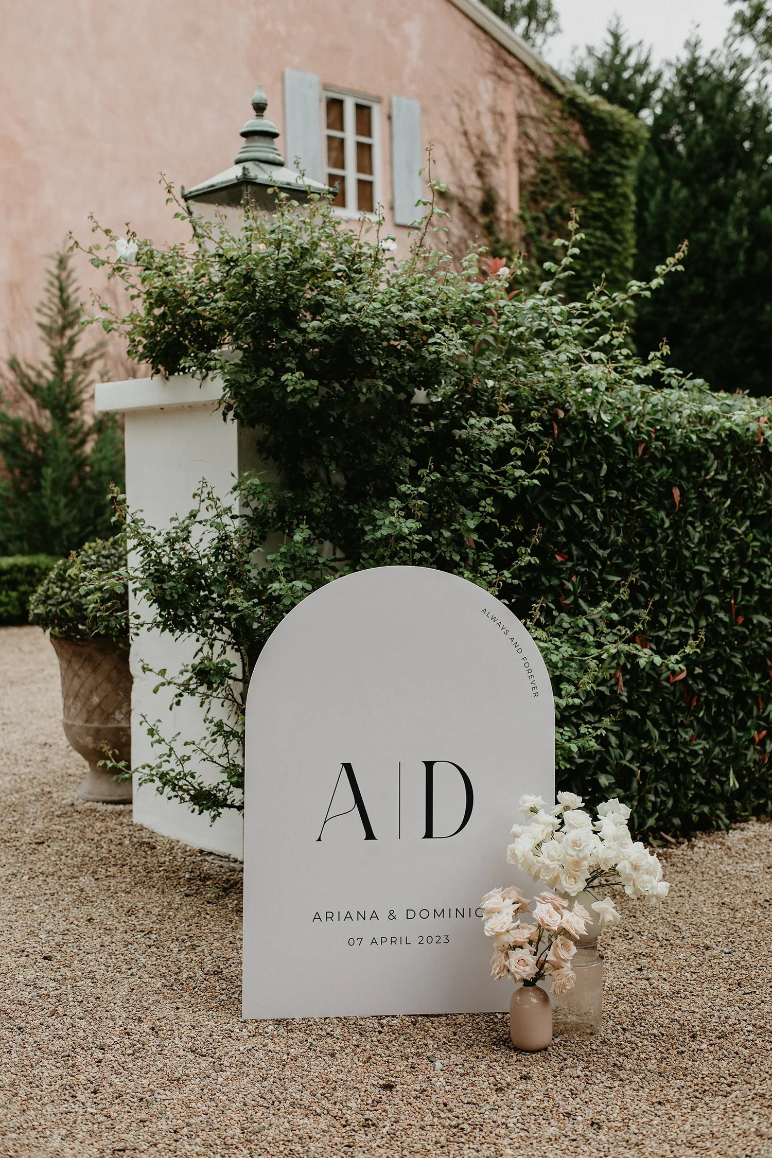 Modern arch-shaped wedding welcome sign for Ariana and Dominic, featuring bold typography and soft floral accents, displayed in a picturesque outdoor venue with a rustic building and lush greenery