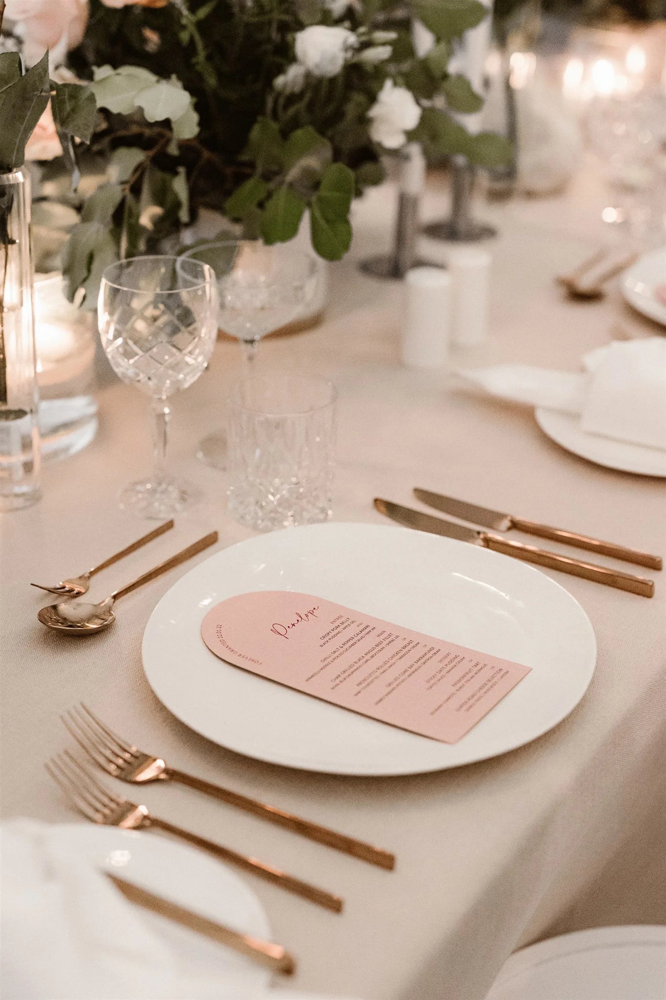 Elegant arch-shaped blush wedding menu card with a printed guest name, displayed on a neutral-toned table setting with gold cutlery and crystal glassware