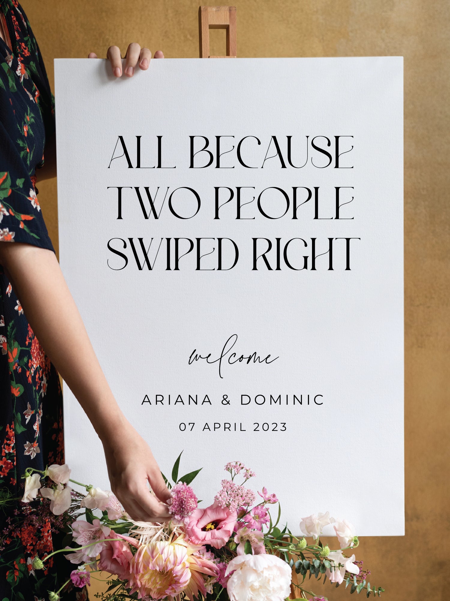  A playful white wedding welcome sign that reads “All Because Two People Swiped Right” in bold black serif font. The couple's names, Ariana and Dominic, along with the wedding date, are displayed below. A floral arrangement with pink, white, and green blooms decorates the easel.

