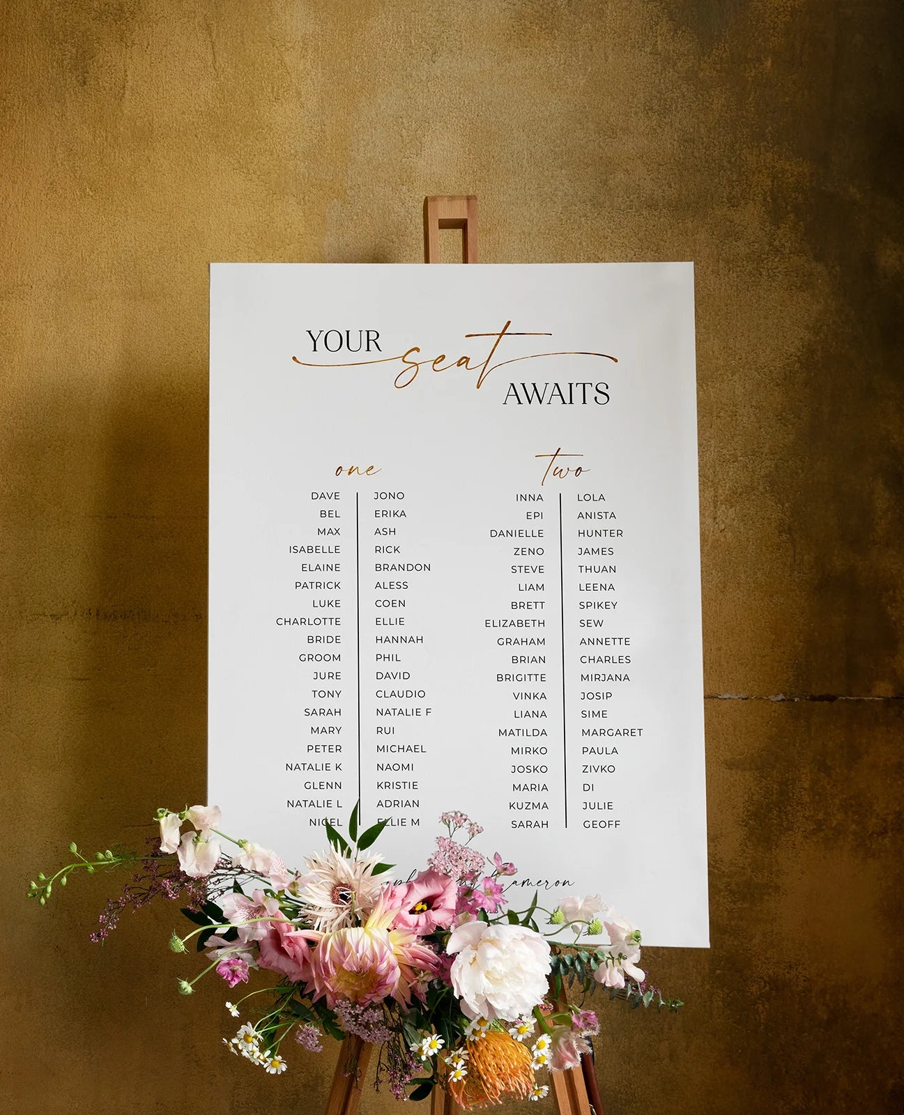 A minimalist wedding seating chart on a white board, with “Your Seat Awaits” in gold script at the top. Guest names are neatly arranged in two columns under table numbers, with a colourful floral arrangement placed at the base of the easel.