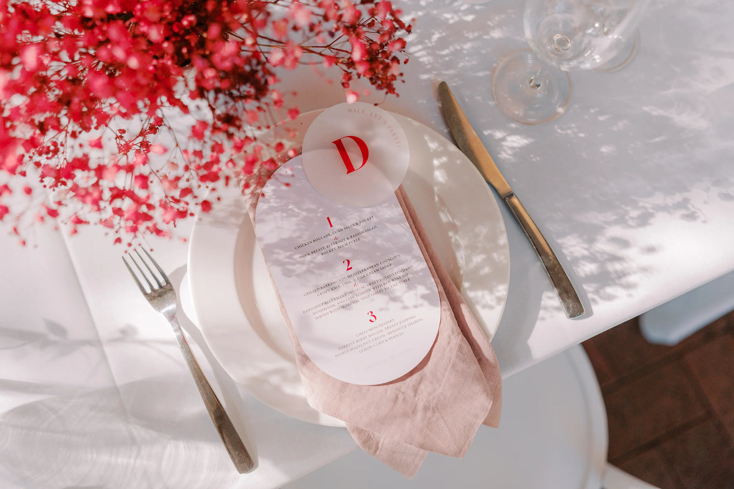 Wedding table setting featuring a double-arch blush menu with red accents, topped with a round acrylic coaster printed with a bold red initial.