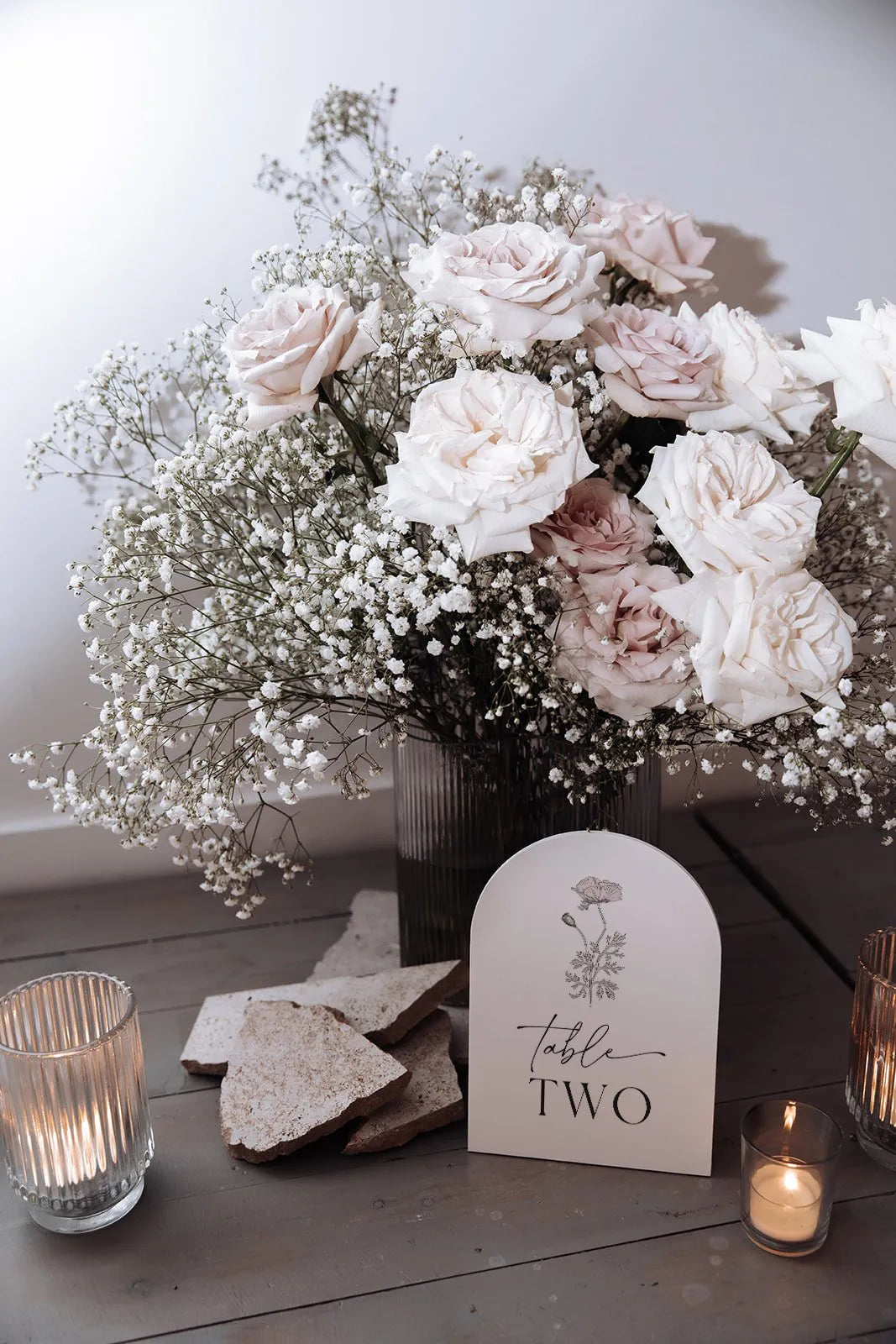 Elegant wedding table number sign in arched shape, surrounded by soft pink roses, baby's breath, and candlelight for a romantic setting