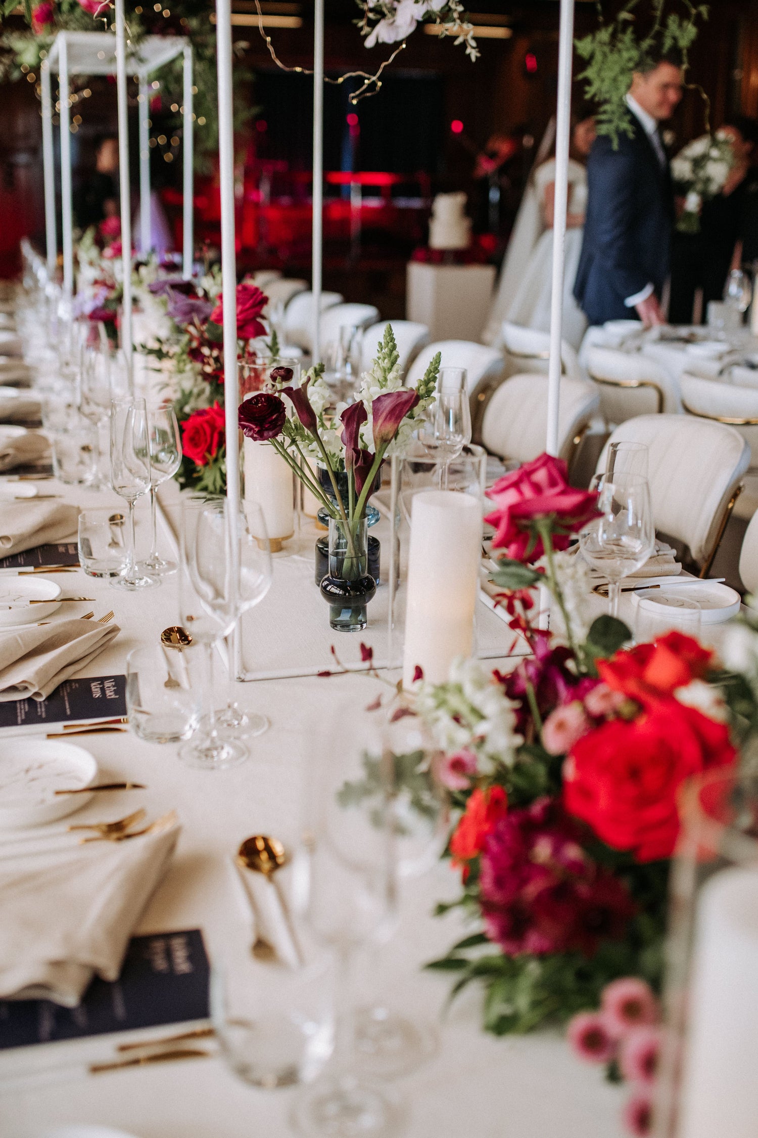 Long wedding table setup featuring vibrant floral arrangements with red, purple, and white flowers, accented by tall candles, glassware, and elegant gold cutlery.