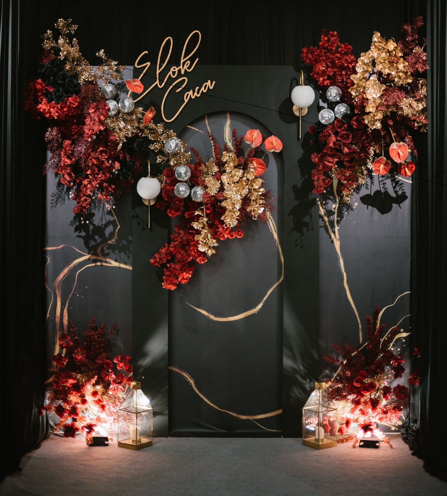 Elegant wedding backdrop with a black arch adorned with red and gold floral arrangements, glowing lanterns, and a personalised sign in cursive lettering.