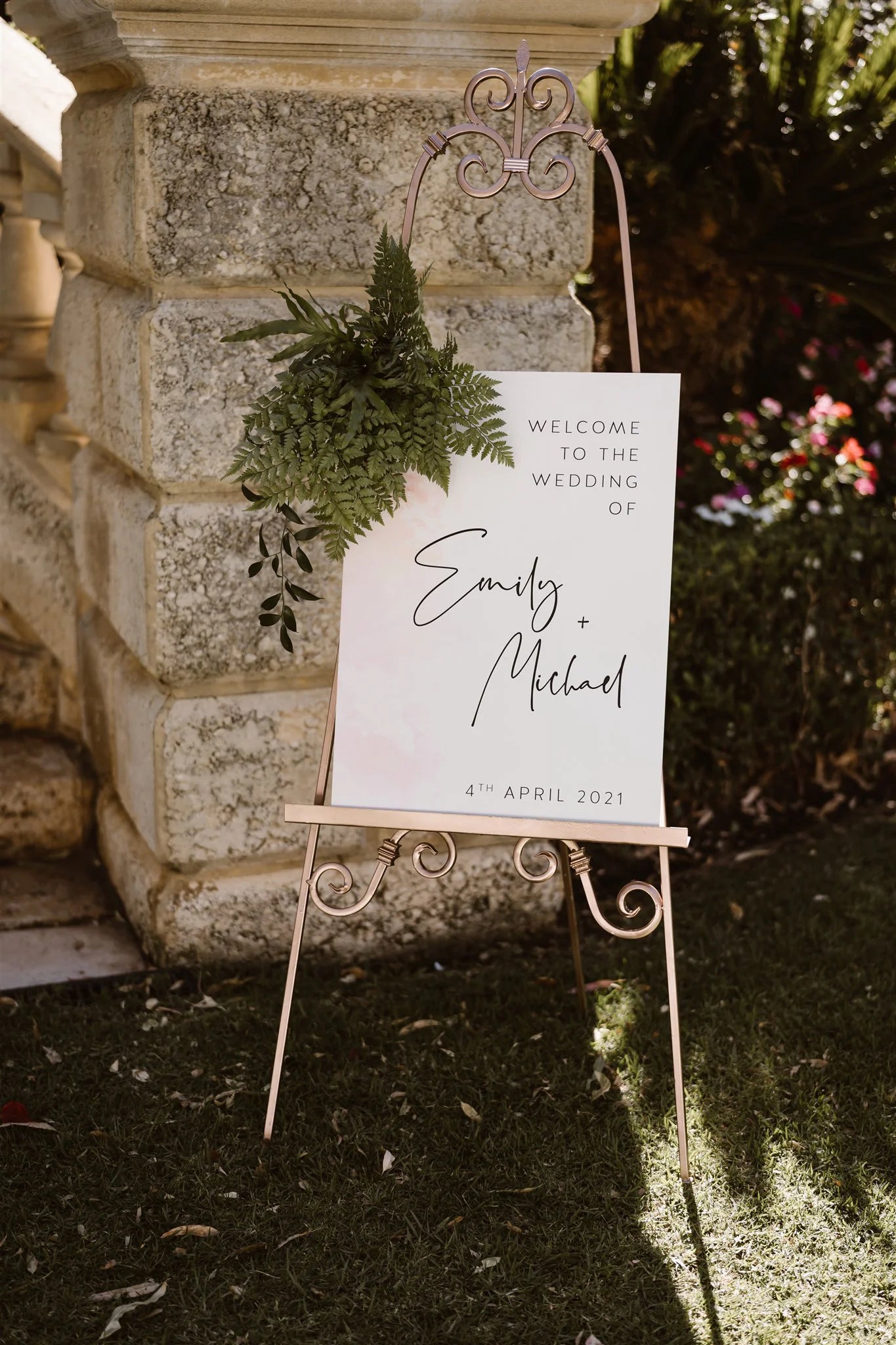 Outdoor wedding welcome sign for Emily and Michael, placed on a vintage wrought-iron easel with greenery accents against a rustic stone wall and lush garden backdrop