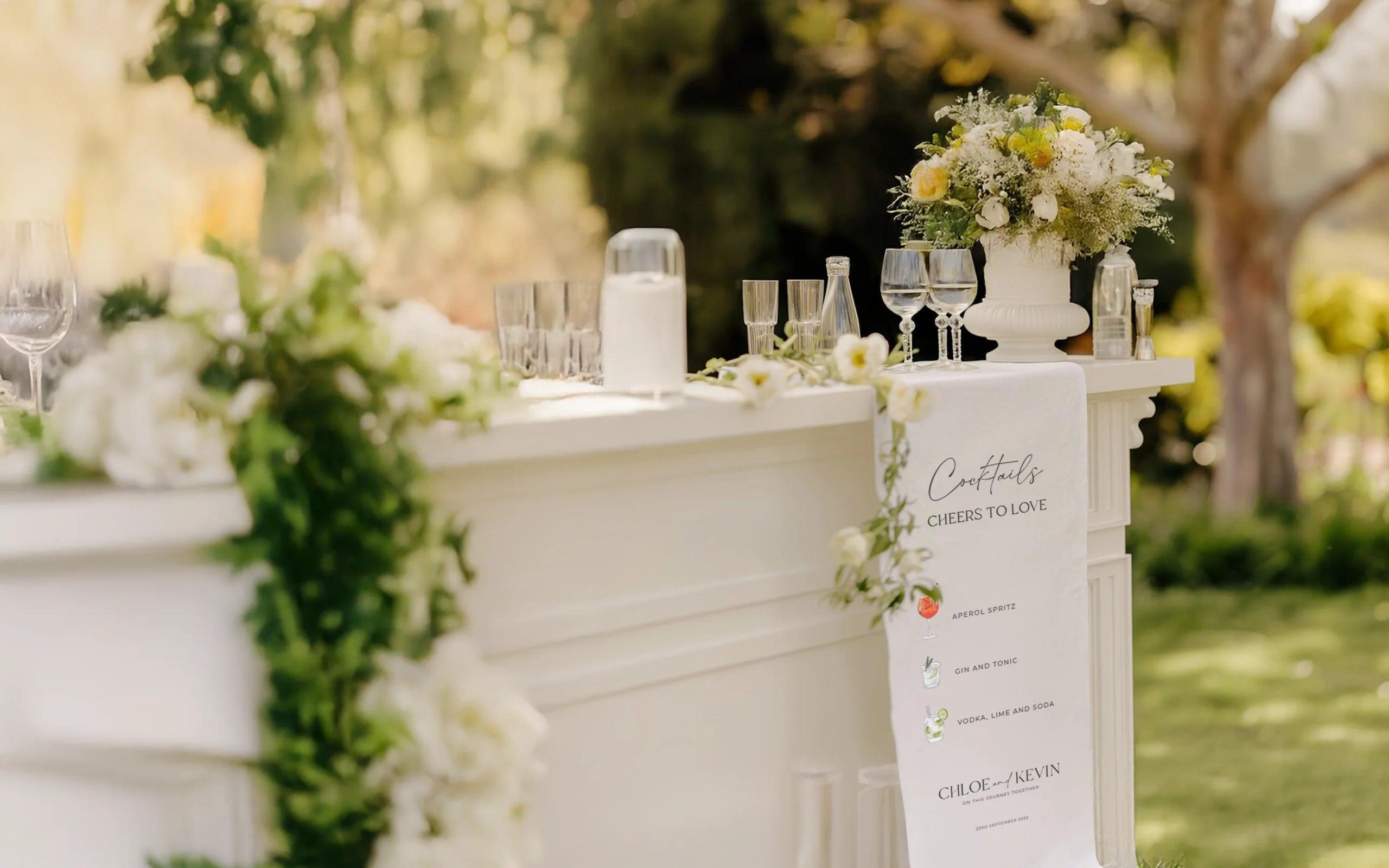 Elegant white wedding bar decorated with green foliage, white flowers, and a floral centerpiece, featuring a cocktail menu banner with 'Cheers to Love' and drink options.