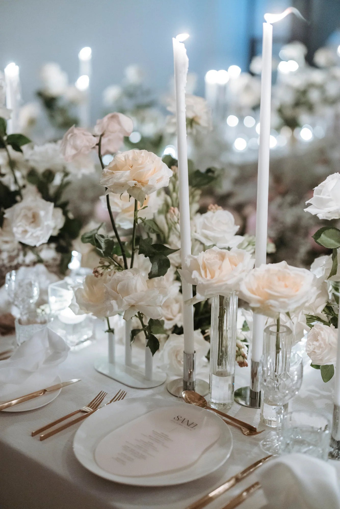 Elegant wedding table setting with tall white candles, ivory roses in glass vases, crystal glassware, and a white menu card on a dinner plate