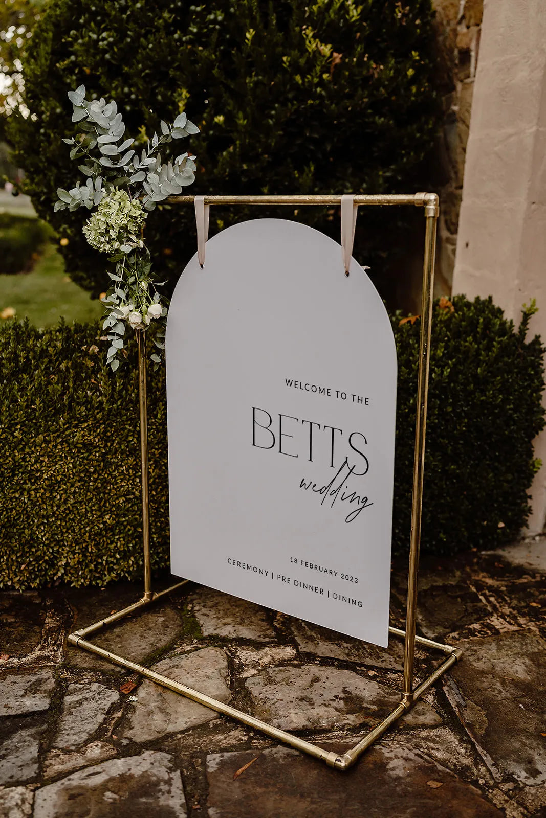 A modern, arch-shaped white wedding welcome sign with “Welcome to the Betts Wedding” written in black script and serif fonts. The sign hangs from a gold frame adorned with eucalyptus leaves and white floral accents.