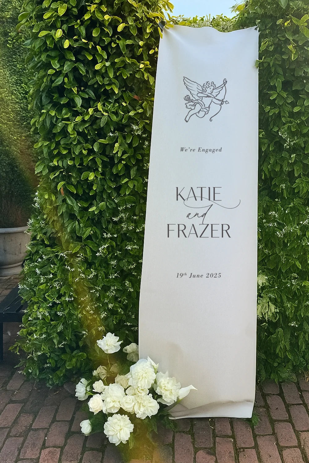 Engagement announcement fabric banner with floral decorations against a green hedge.