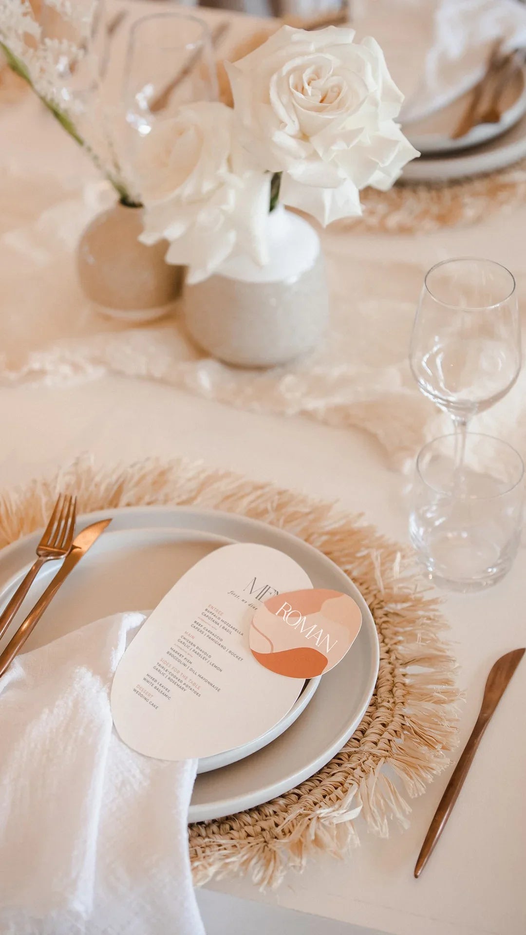 Elegant wedding table setting featuring an arch-shaped menu card with soft blush tones and a matching round place card with the guest's name, displayed on a neutral plate with woven placemat, accompanied by white roses in minimalist vases.