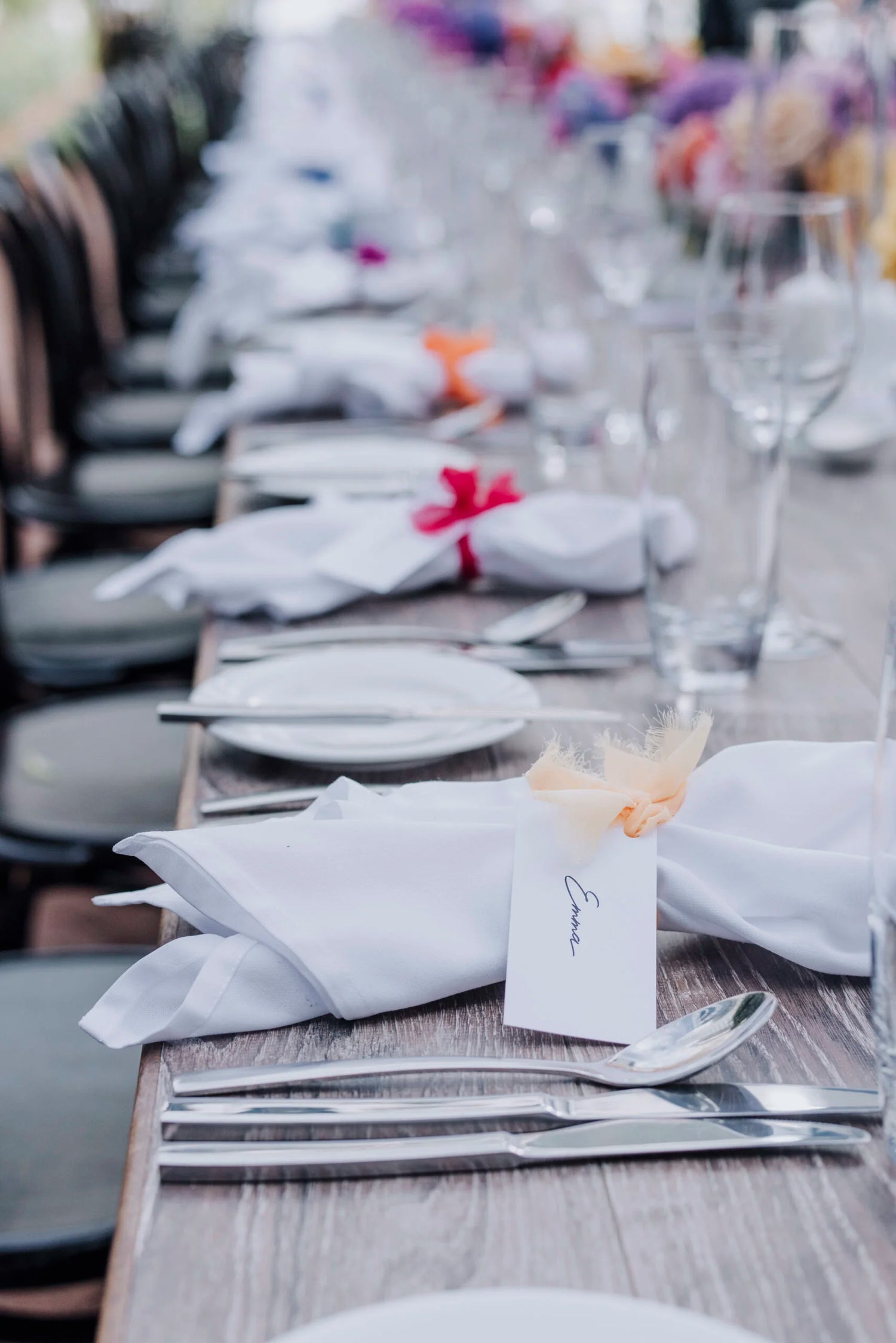 Close-up of wedding place settings featuring neatly folded white napkins tied with colourful ribbons, personalised guest name cards, and polished silver cutlery on a rustic wooden table