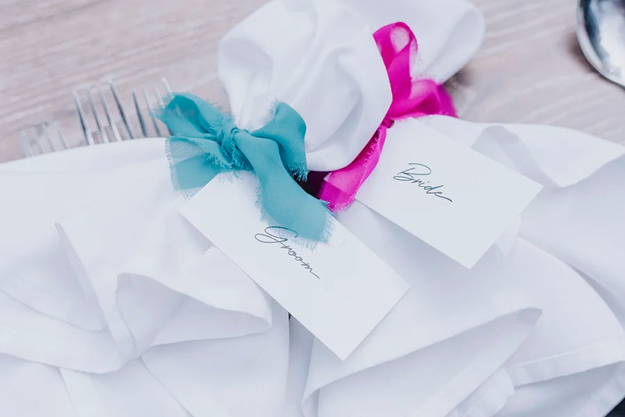 Close-up of wedding place settings featuring white napkins tied with colorful ribbons, personalised guest name cards, and polished silver cutlery on a rustic wooden table