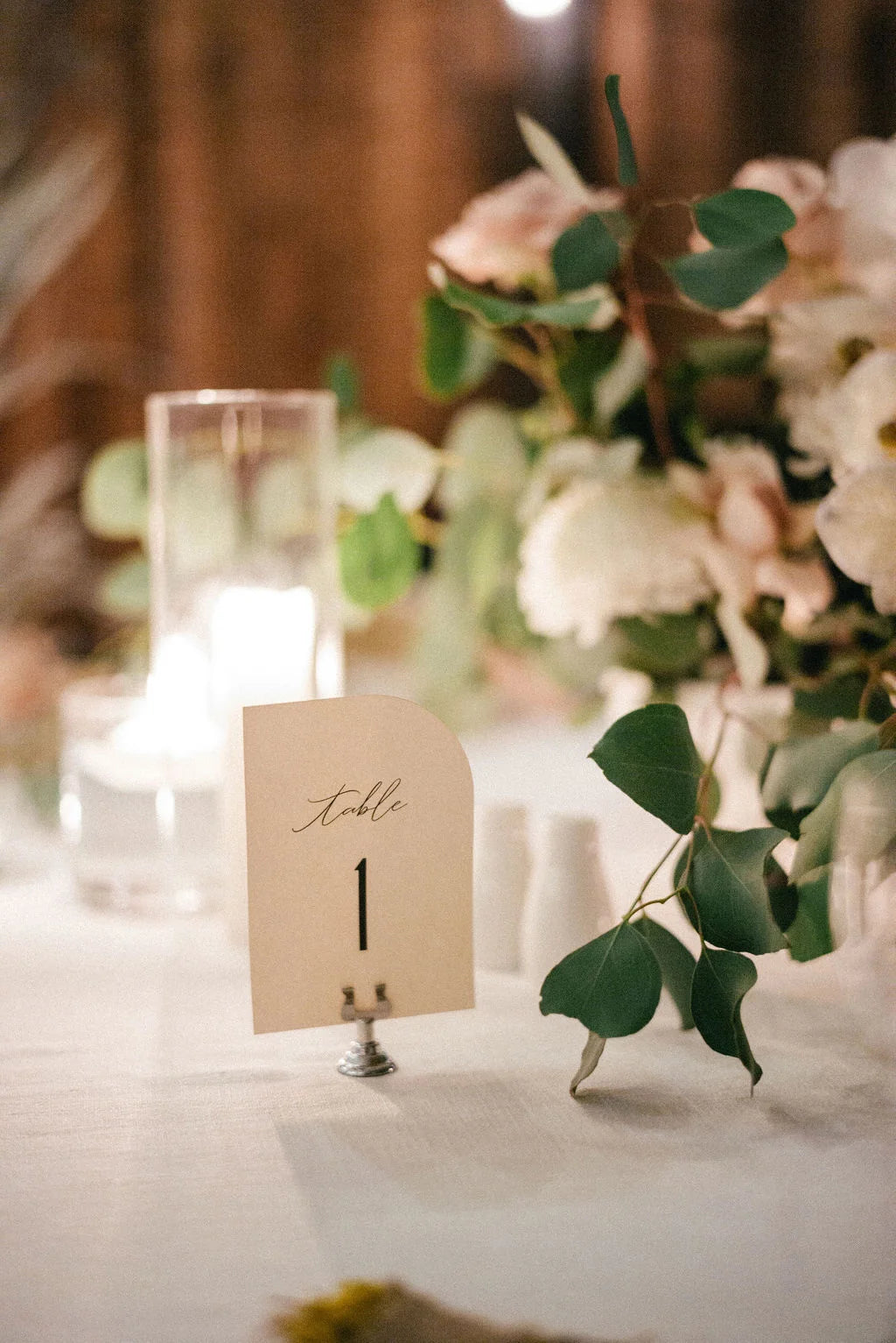 Minimalist cream sail-shaped table number card displaying “Table 1,” styled among soft candlelight and lush floral arrangements.