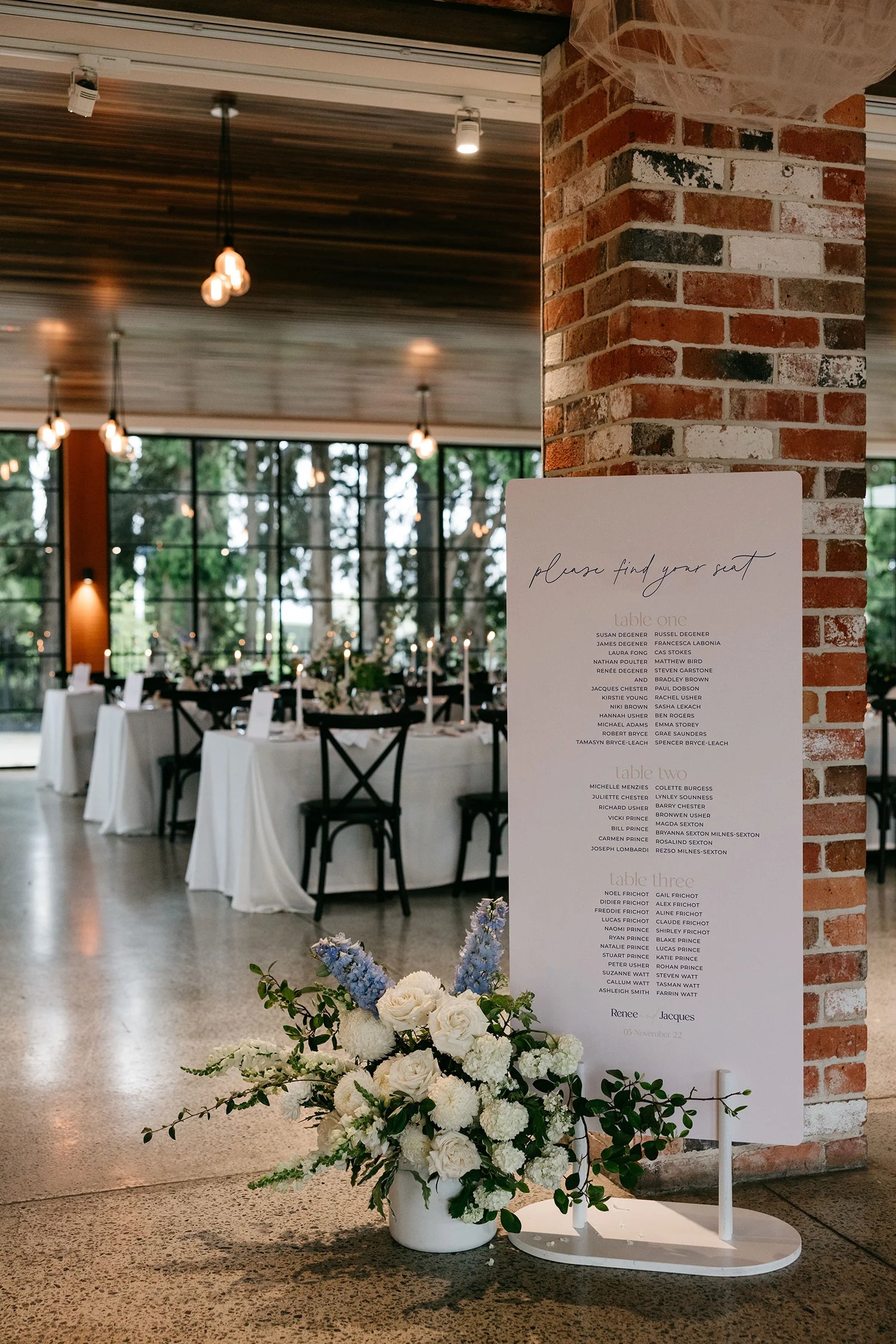 A sleek wedding seating chart is displayed at the base of a rustic red brick pillar inside a wedding reception venue. The seating chart, titled “Please Find Your Seat,” lists guests’ table assignments in a clean, minimalist font. A floral arrangement with lush white roses and soft blue flowers sits at the base of the sign, complementing the modern white stand. In the background, tables draped in white linens are set for the reception, with black chairs and candles adding to the atmosphere.